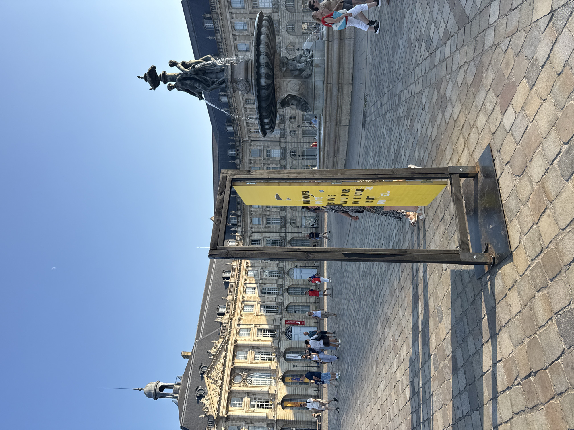 In Bordeaux, France, the Place de la Bourse is depicted on a clear day. The iconic Bourse fountain is visible in the background, with water cascading from a sculptural group. The grand architecture of the Place de la Bourse buildings frames the scene. A yellow sign is prominently positioned in the foreground, supported by a dark metal frame, with text partially visible. Several people are walking across the cobblestone plaza, some appearing to be tourists. The lighting suggests it is late afternoon or early evening, casting long shadows across the ground. The sky is a clear blue with a faint crescent moon visible.