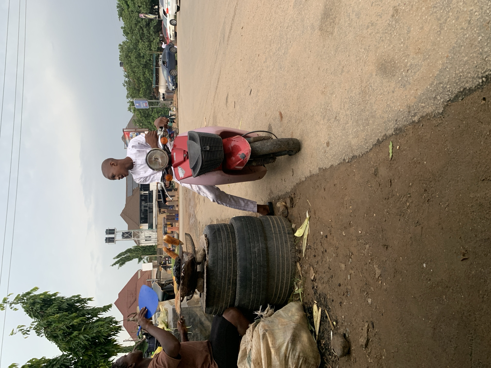 The image depicts a street scene in Abuja, Nigeria, where two individuals are interacting near a road. In the foreground, a person wearing a brown t-shirt and dark shorts is seated, holding a blue object and attending to a grill with corn on it. The grill is elevated by a stack of three tires. To the right, another individual, dressed in a white traditional outfit, is seated on a red and pink scooter. The scooter has a black basket in front. Both individuals appear to be engaged in conversation or a transaction. The environment is dusty, with a paved road on the right and a dirt area on the left. In the background, various buildings, trees, and vehicles are visible, suggesting an urban setting within Abuja. The sky is overcast.