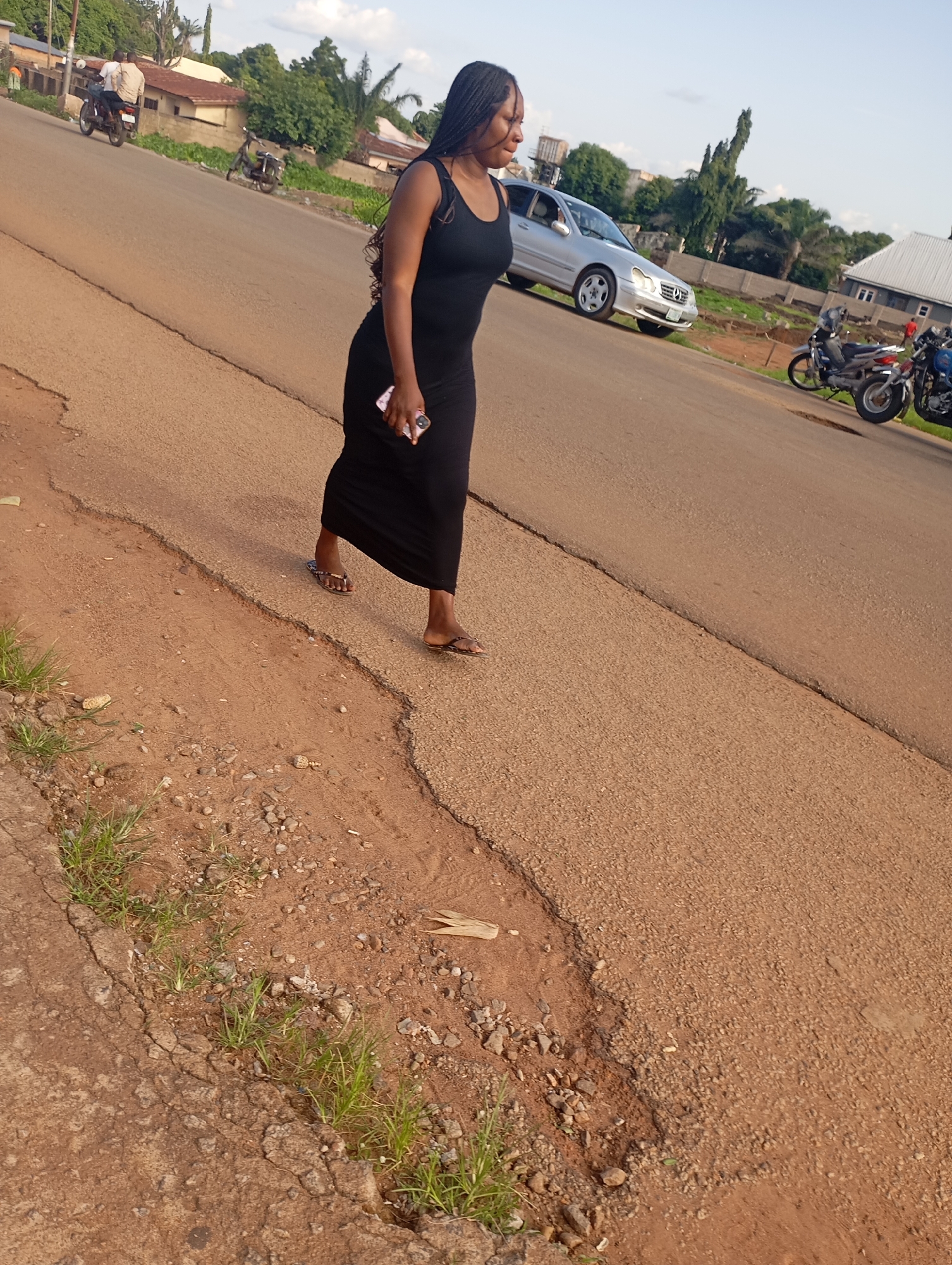 In Shendam, Nigeria, a young woman with braided hair walks along a paved road. She is wearing a long, black dress and holding a pink smartphone in her right hand. She is walking from left to right in the frame, with her body angled towards the viewer. The road is made of asphalt and is in moderate condition, with some cracks and unevenness. To the left of the road, there is a dirt shoulder with patches of grass growing through it. In the background, there are several buildings, including houses and what appears to be a small commercial establishment. There are also several motorcycles parked on the side of the road, and a silver car is visible further down the road. The sky is blue with some scattered clouds, and the overall lighting suggests it is late afternoon.