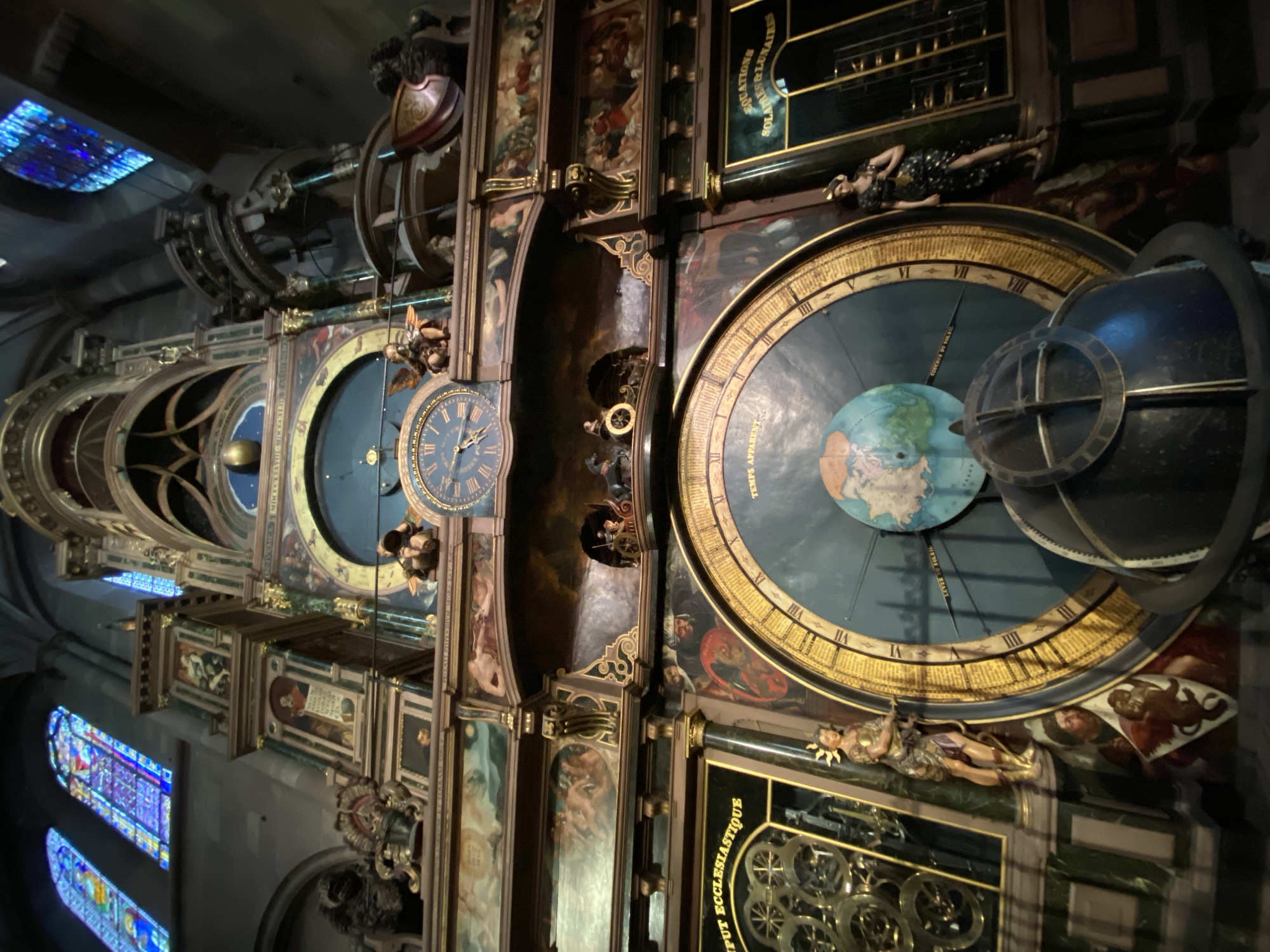 This image captures a detail of a large, ornate astronomical clock, likely located within a grand cathedral or historical building. The clock is intricately designed with multiple dials, figures, and decorative panels, all appearing to be made of wood, brass, and painted elements, with gilded accents. The overall aesthetic is rich and complex, indicative of historical craftsmanship.
The main subject is the clock itself, which features several faces. On the right, a prominent circular dial displays Roman numerals from III to XII (clockwise). This dial incorporates a terrestrial globe depicting landmasses, and another, larger celestial globe or armillary sphere. Text visible on this dial includes "TEMPS APPARENT" (Apparent Time) and partially visible "LEVER DE SOLEIL" (Sunrise). Below this main dial, a panel with golden lettering on a dark background reads "COMPUT ECCLESIASTIQUE" (Ecclesiastical Computus), referring to calculations for the church calendar. Above it, another dark panel with gold lettering includes "SOLUTIONS LUNAIRES" (Lunar Solutions/Calculations).
To the left, another smaller clock face with Roman numerals is visible. The clock structure is adorned with various figurines, including a reclining female figure with a star-patterned dress near the upper right dial, and a golden figure with sunrays around its head near the lower right. Painted panels with allegorical scenes or historical figures decorate the vertical sections between the dials. Some intricate brass gears and mechanical components are visible in the lower right, part of the clock's complex inner workings.
In the background and to the left, several vibrant stained-glass windows cast colorful light, indicating the interior setting is a grand edifice. The lighting within the space appears dim, enhancing the historical and artistic quality of the clock. No specific human activity is depicted other than the implicit function of the clock to measure and display time and astronomical phenomena. The image is rotated 90 degrees clockwise. This object is consistent with the famous astronomical clock found in Strasbourg Cathedral.