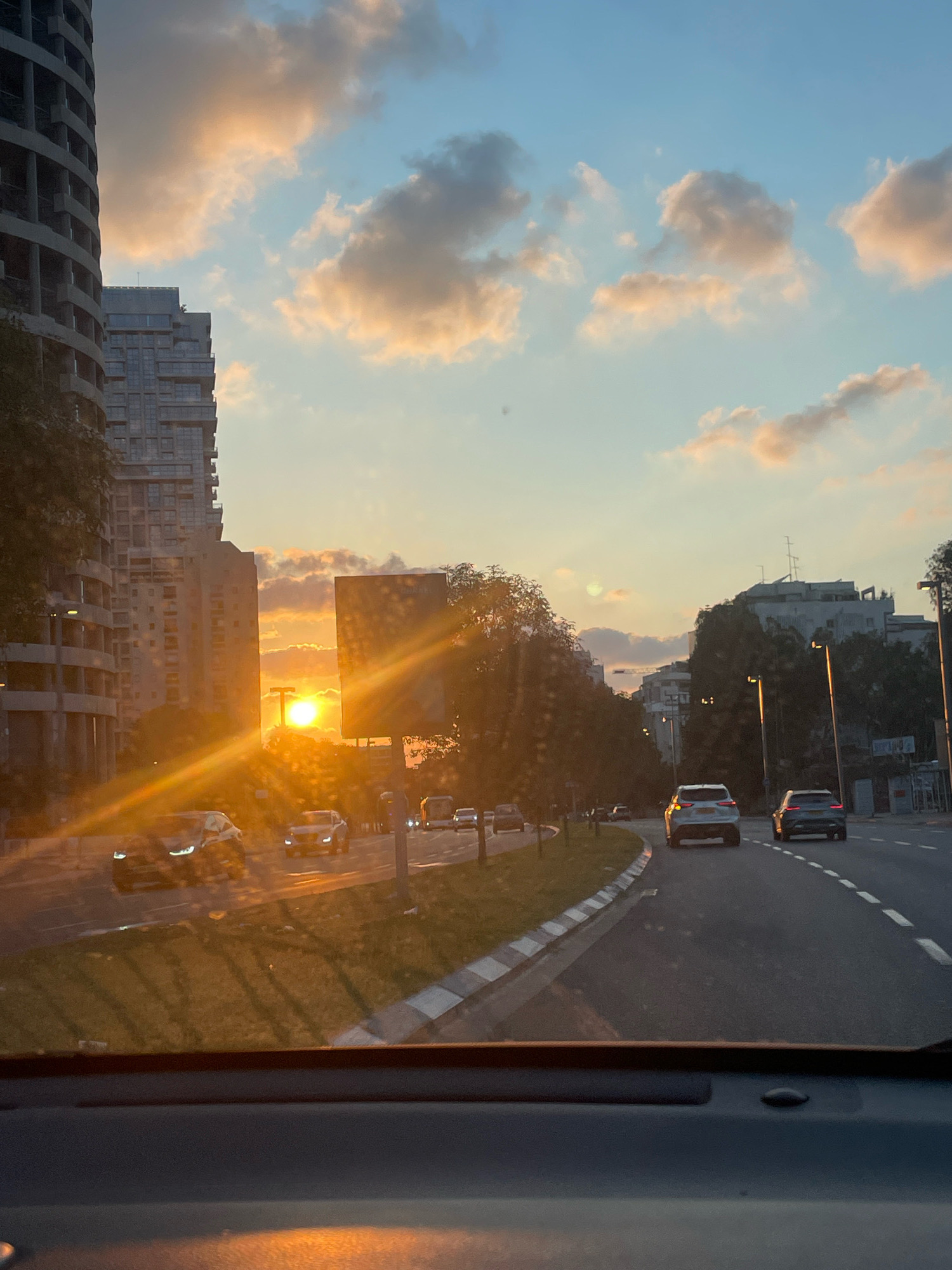 The image captures a street scene in Tel Aviv, Israel, during a sunset. The sun, a bright orb, is positioned low on the horizon, casting long golden rays across the sky and the road. The sky is a blend of soft blues and warm oranges, with scattered clouds tinged with the sunset's light. On the left side of the frame, modern high-rise buildings dominate the cityscape, their concrete facades reflecting the ambient light. A row of trees lines the median of the multi-lane road, with streetlights visible among them. Several cars are on the road, moving in the same direction, with headlights of some vehicles illuminated. The perspective is from inside a car, looking out through the windshield, which shows some condensation or dust particles. The overall impression is a typical urban evening commute with the added beauty of a vibrant sunset.