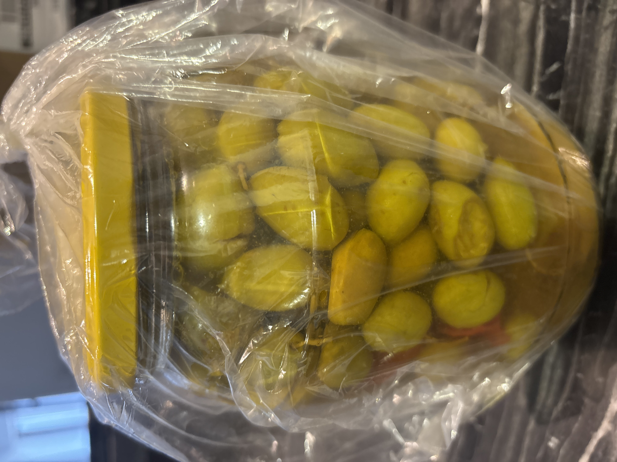 A close-up, eye-level shot shows a glass jar filled with green olives, submerged in oil. The jar has a yellow lid and is partially covered by a clear plastic bag. The olives are plump and vary in shape, some appearing smooth and others with slight indentations. A few slivers of red, possibly chili peppers, are visible amongst the olives at the bottom of the jar. The jar rests on a dark, patterned surface, possibly a countertop or table. The background is blurred, showing hints of vertical blinds or window coverings. The lighting is overhead, casting soft shadows. No people or animals are present in the image. The scene is indoors and appears to be in a kitchen or pantry. No specific location in Dubai, United Arab Emirates, can be determined from the visual information provided.