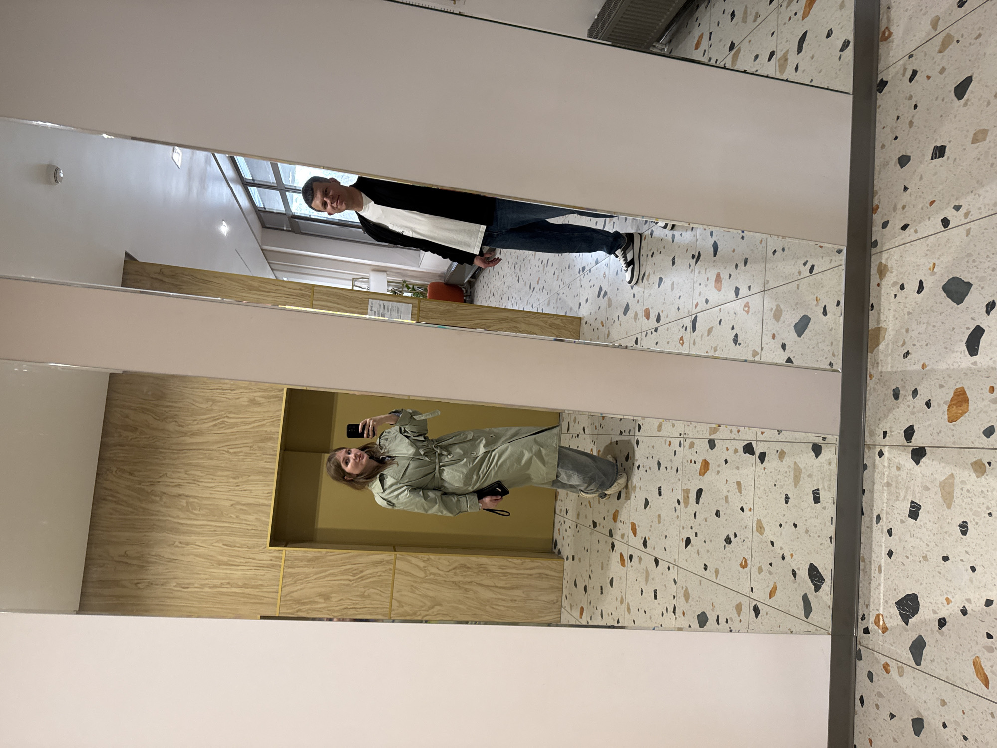 The image shows a mirrored reflection of two individuals and the surrounding environment, possibly within a building in Perm, Russia, based on the URL and the general aesthetic. A woman is positioned near the mirror, facing it, with a smartphone in her hand, presumably taking a selfie. She is wearing a light green trench coat and jeans. A man stands behind her in the reflection, further away from the mirror. He is wearing a white shirt, black jacket, and jeans. The floor features a terrazzo tile pattern with a mix of colors including black, orange, and grey. The wall behind the woman appears to be covered in wood paneling with a yellow trim. The ceiling is white and includes a ventilation grate.