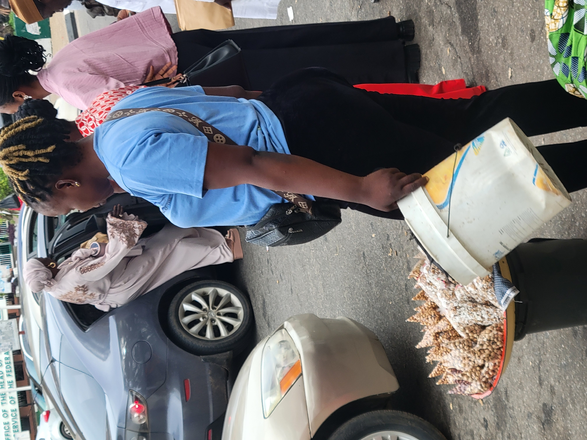 The vertically oriented image captures an outdoor street or market scene, likely in Abuja, Nigeria.
In the foreground, a woman with braided hair featuring blonde highlights, dressed in a light blue t-shirt and black trousers, is prominent. She wears a black shoulder bag with a distinct brown, patterned strap. She is actively holding and tilting a large white plastic bucket over a wide, reddish-orange basin resting on the ground. The basin is meticulously filled with numerous small, conical-shaped, light-brown items, possibly individually packaged groundnuts or a local snack, arranged in neat rows for sale or display. A black bucket sits next to the basin.
Behind this woman, another woman is partially visible, wearing a pink top over a red and white patterned blouse and black trousers. Further back, glimpses of other people, including one in a light brown cap, suggest a bustling environment.
To the left of the frame, a dark grey sedan is parked. A person, possibly a woman, adorned in a light brown or mauve long dress with embroidery and a matching head covering, is seen leaning out of the car's window. The front portion of a beige sedan is also visible in the lower right.
A white sign or plaque on a building or wall near the dark grey car is partially legible, displaying text that reads "OFFICE OF THE HEAD OF..." and "...SERVICE OF THE FEDERAL...", indicating a government or public service establishment. The scene takes place during daytime, under natural light, on a paved surface. The activity suggests a typical urban setting where goods are being sold or displayed.