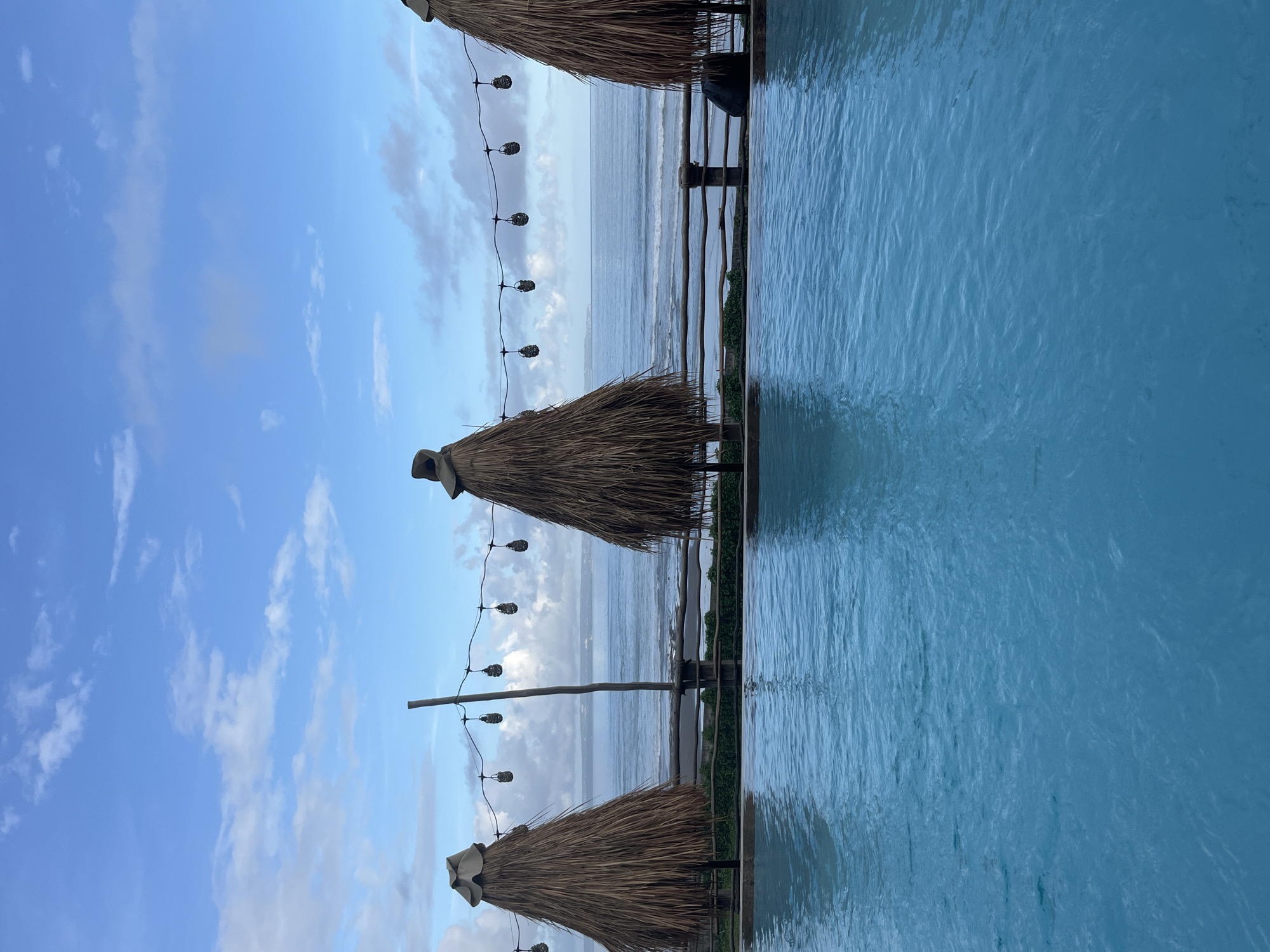 This is an eye-level, outdoor shot in Mengwi, Indonesia, featuring three thatched-roof umbrellas on a wooden deck beside a swimming pool. A string of lights is strung between the umbrellas. In the background, the ocean meets a cloudy sky.