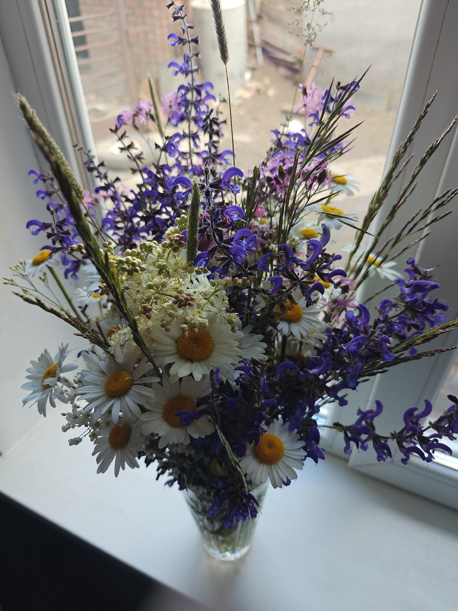 A vase of wildflowers is on a windowsill in Knyshivka, Ukraine. The bouquet contains daisies, purple and pink wildflowers, and grasses. The vase is transparent and filled with water, and the windowsill is white. The window looks out onto a blurred cityscape.