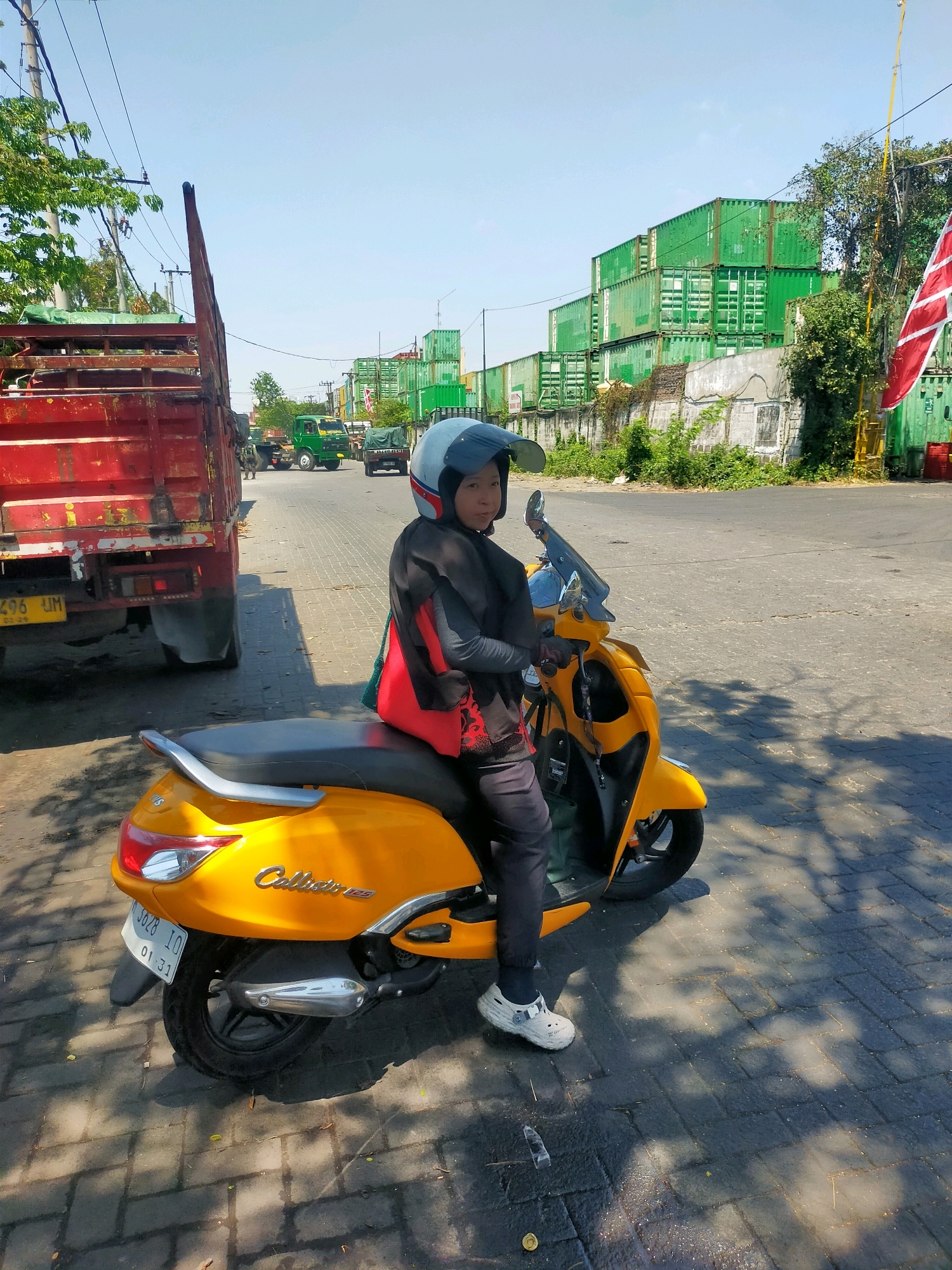 A child wearing a helmet sits on a yellow scooter in an industrial area in Surabaya, Indonesia. The child is wearing a black jacket with red accents and black pants, with white shoes. The scooter has a license plate that reads "1628 IQ 01-28". In the background, there are stacks of green shipping containers, a red truck, and other vehicles. The ground is paved with brick. The sky is clear and blue.