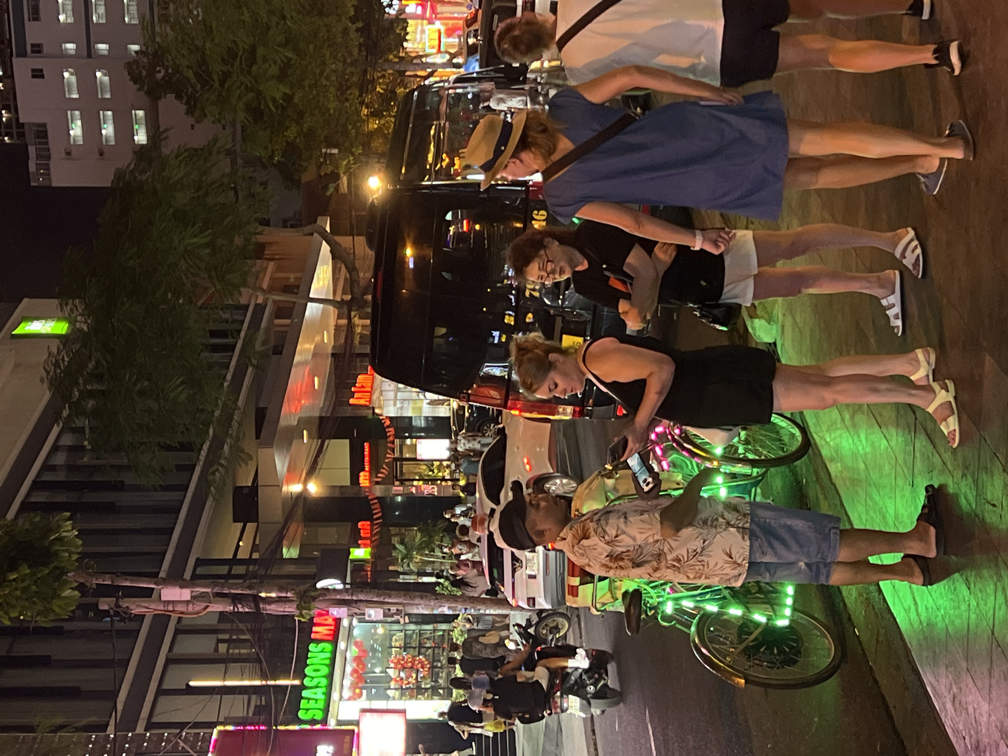 The image shows a group of six people standing on a sidewalk at night, next to bicycles with green lights, in Nha Trang Ward, Vietnam. The street scene includes vehicles, lit-up storefronts and signs ("SEASONS MAY" visible) and street lights. One person is looking at a phone, while others are looking in different directions.