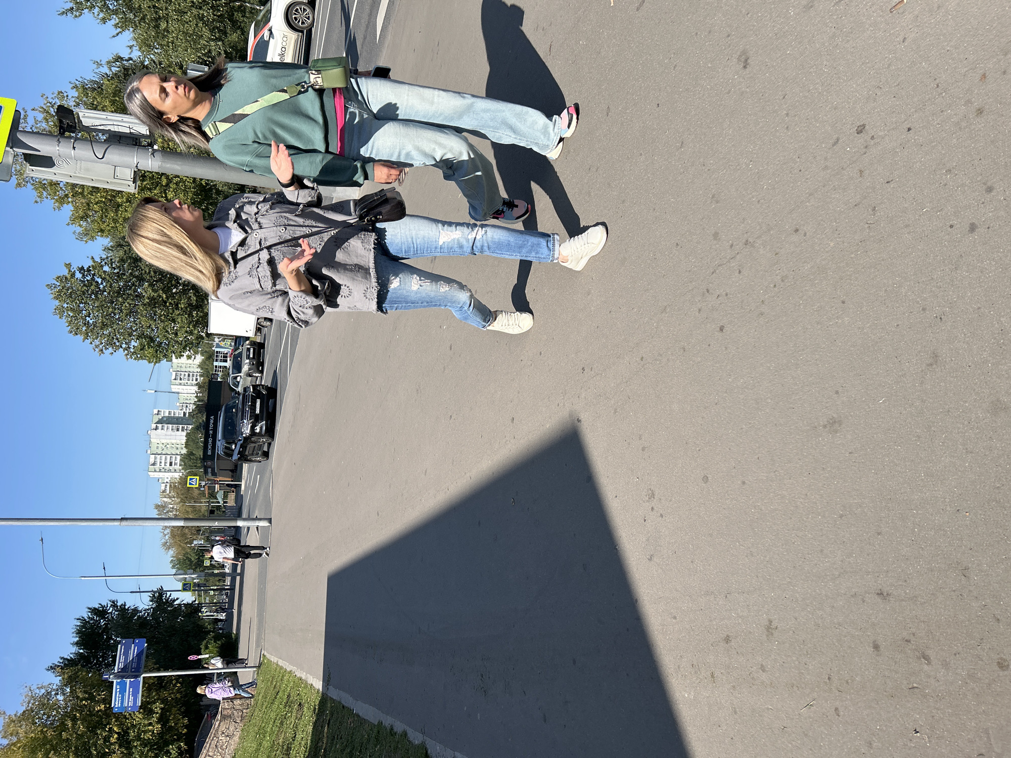 Two women walk on a sidewalk in Moscow, Russia, under a clear blue sky. The woman on the left, with blonde hair, wears a grey patterned jacket, a white t-shirt, and ripped blue jeans with white sneakers. She carries a black crossbody bag and gestures with her left hand. The woman on the right, with grey-streaked blonde hair, wears a green sweatshirt, light blue jeans, and multi-colored sneakers. She has a green crossbody bag and a pink belt visible under her sweatshirt. They are walking away from the viewer, towards a street with cars, including a black SUV and a white car. The sidewalk is paved, with a grassy embankment to the left and a tall lamppost casting a long shadow. Traffic signs and tall residential buildings are visible in the background.