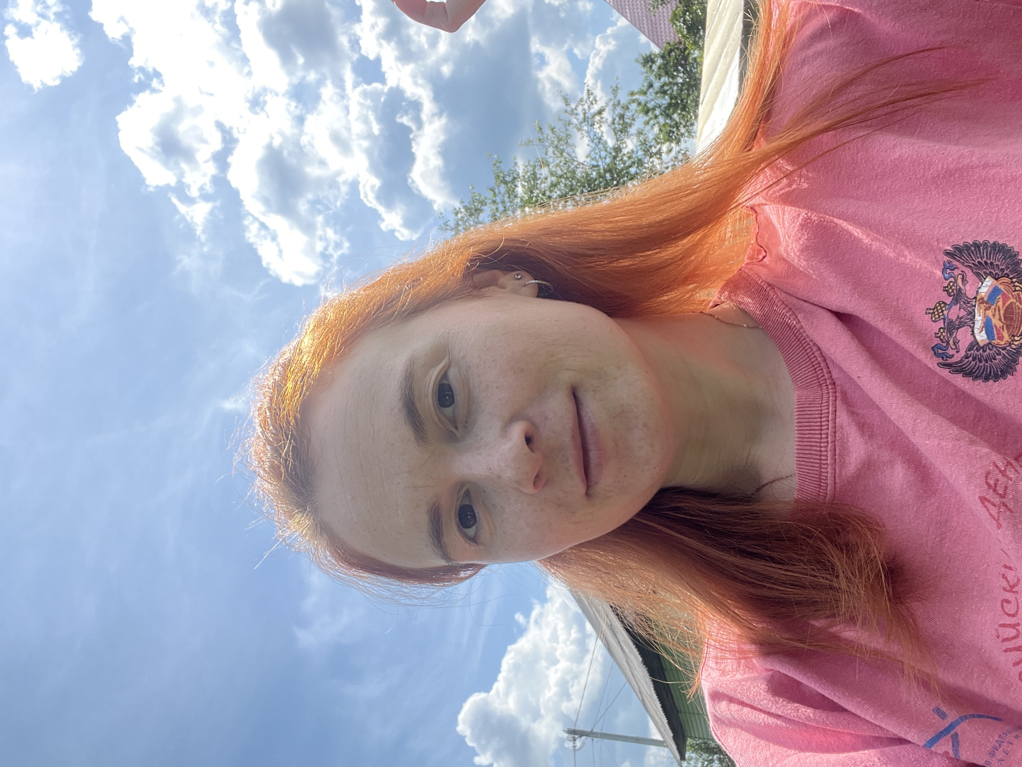 A selfie taken outdoors on a sunny day shows a woman with vibrant red hair and freckles looking at the camera. She is wearing a pink sweatshirt. Behind her, the sky is blue with white, puffy clouds. The background also features trees, a green building with a grey roof, and a glimpse of another building with a red roof. The location is the city of Igra, Russia.