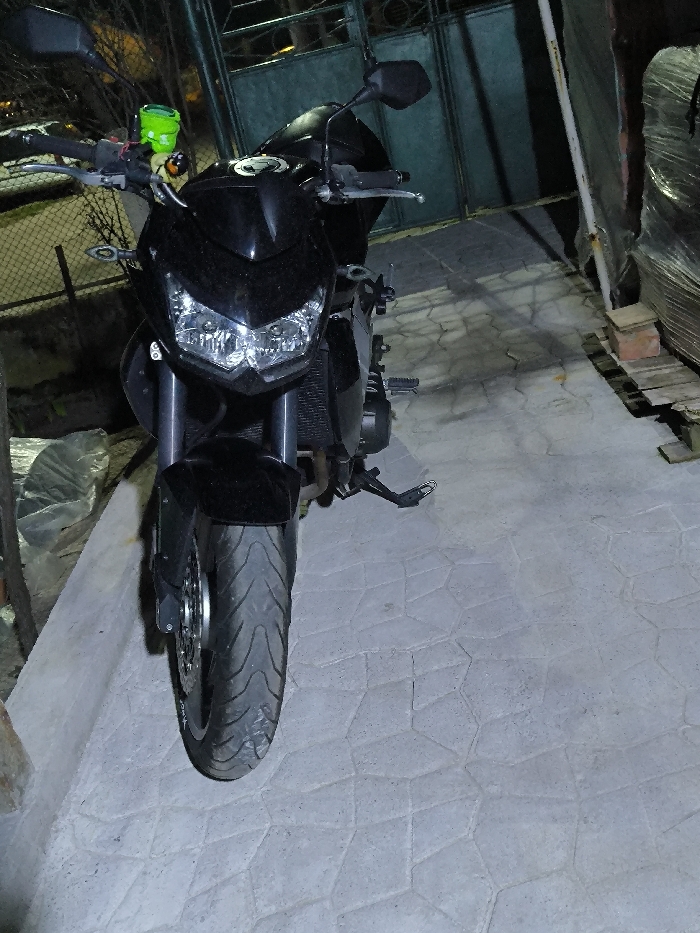 A black motorcycle is parked facing forward on a light-grey, polygonal-patterned paved surface in Dolistovo, Bulgaria. The motorcycle features dual illuminated headlights, a visible radiator, black handlebars with rearview mirrors, and a distinct neon-green cylindrical fluid reservoir mounted on the left handlebar. The front tire displays a noticeable tread pattern. In the left background, a section of metal mesh fencing is partially visible. In the right background, a dark metal gate is present, and further to the right, a stack of items, possibly wooden pallets, wrapped in clear plastic, rests on the ground, supported in part by a red brick. The scene is illuminated by artificial light.