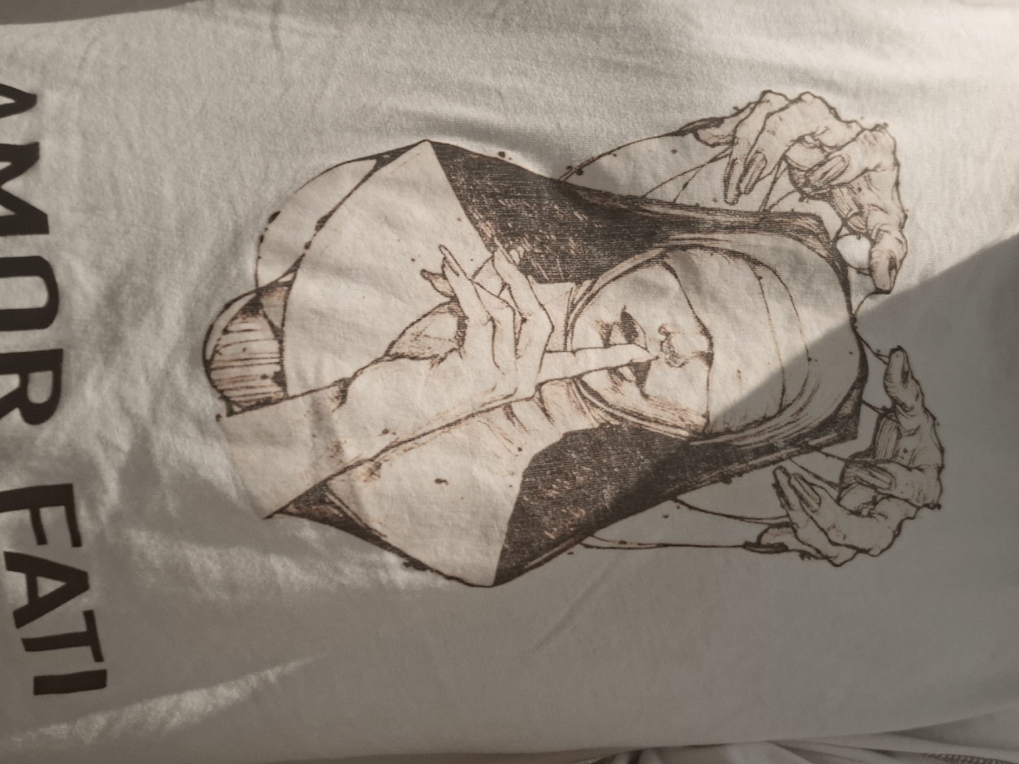 The image depicts a white fabric, likely a shirt, with a dark brown graphic print. The print features a stylized image of a nun with hands gesturing near her face. To the left of the graphic, black text reads "AMOR FATI" vertically. The lighting is warm, creating shadows across the fabric and highlighting the texture of the material. The environment appears to be indoors with the fabric lying flat on a surface. Burgas, Bulgaria is not present in the image.