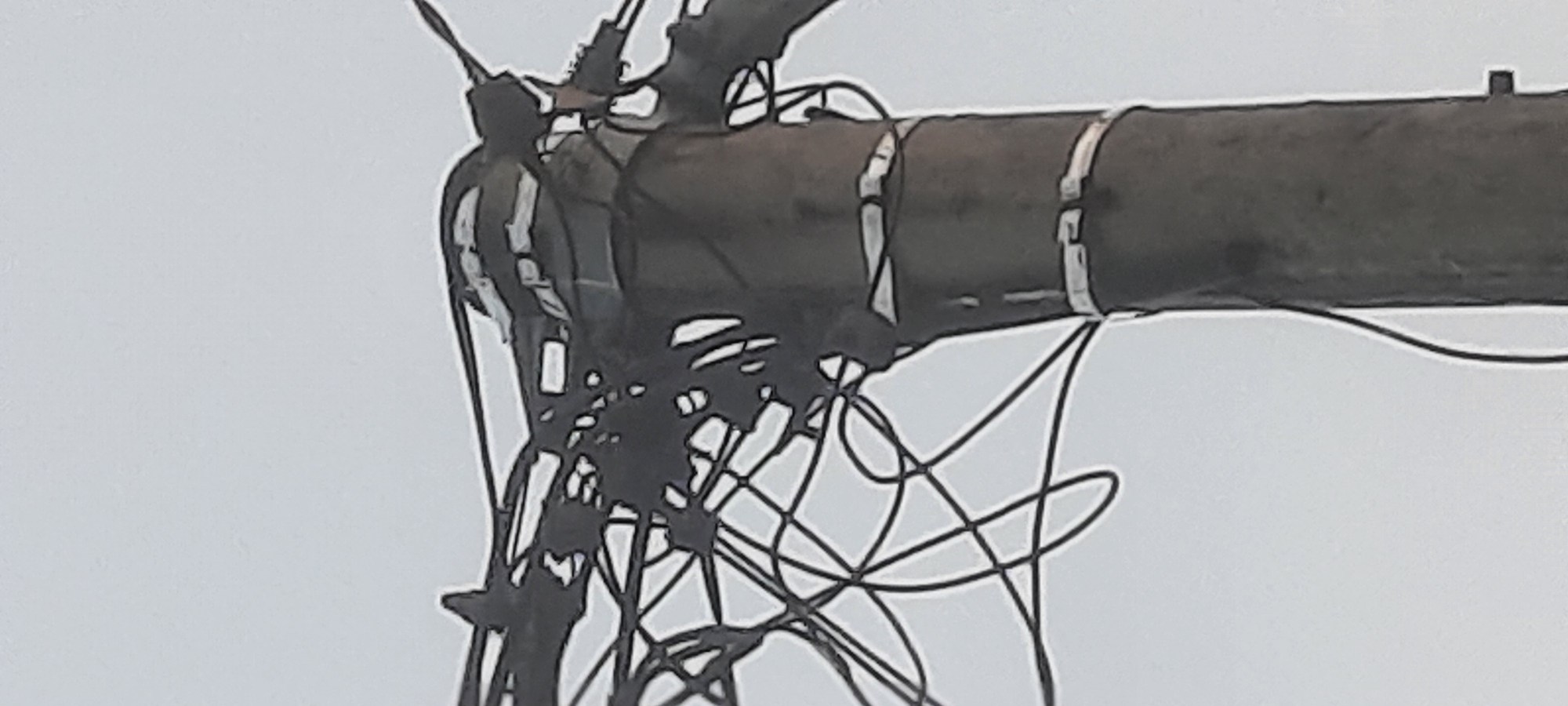 The image shows a close-up view of a utility pole in Ploso, Indonesia, against a clear sky. The pole is made of dark material, possibly concrete or wood, and is wrapped with several white bands at intervals. A thick bundle of electrical wires is attached to the top of the pole, with numerous thin wires extending from it and crisscrossing in front of the pole. Some of the wires appear to be damaged or tangled. The overall impression is one of aged or poorly maintained electrical infrastructure.