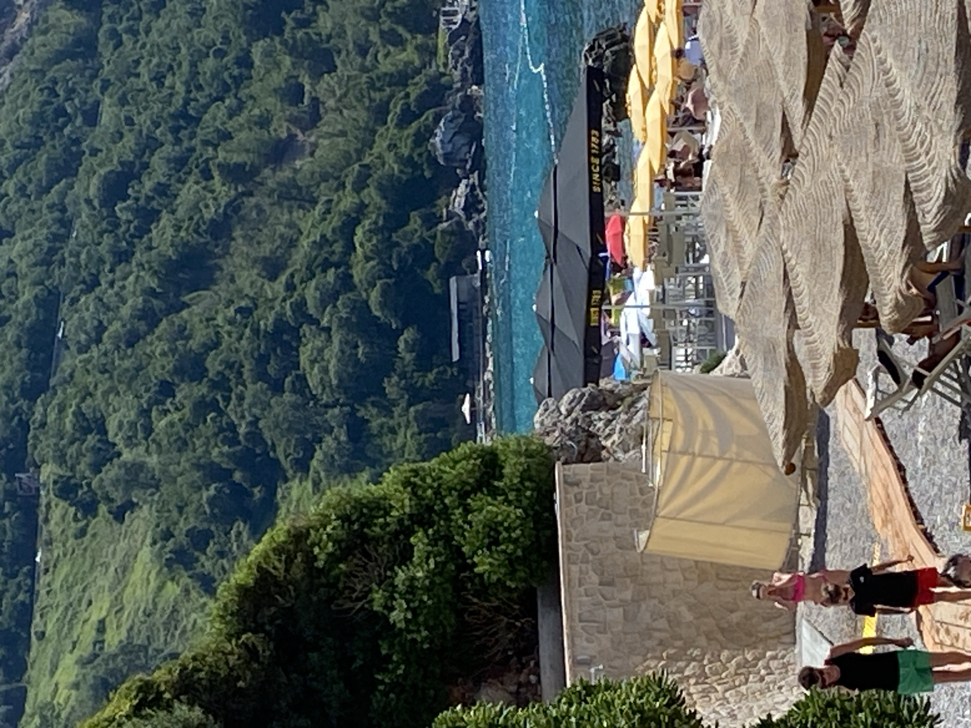 The image depicts a crowded beach scene on a sunny day, with a lush green hillside dominating the background. In the foreground, a wooden walkway leads towards the water, with numerous beach umbrellas providing shade for beachgoers. Some individuals are visible walking along the walkway and relaxing under the umbrellas. A stone wall with a curved, beige curtained structure stands to the left of the walkway. The sea is a vibrant blue, with some white buoys or markers visible in the water. On the hillside, trees and vegetation cover the landscape, and a dark structure, possibly a building or a covered seating area, is visible near the water's edge. In the distance, along the hilly terrain, there appears to be a road or path with vehicles. The location is identified as Prijevor, Montenegro.