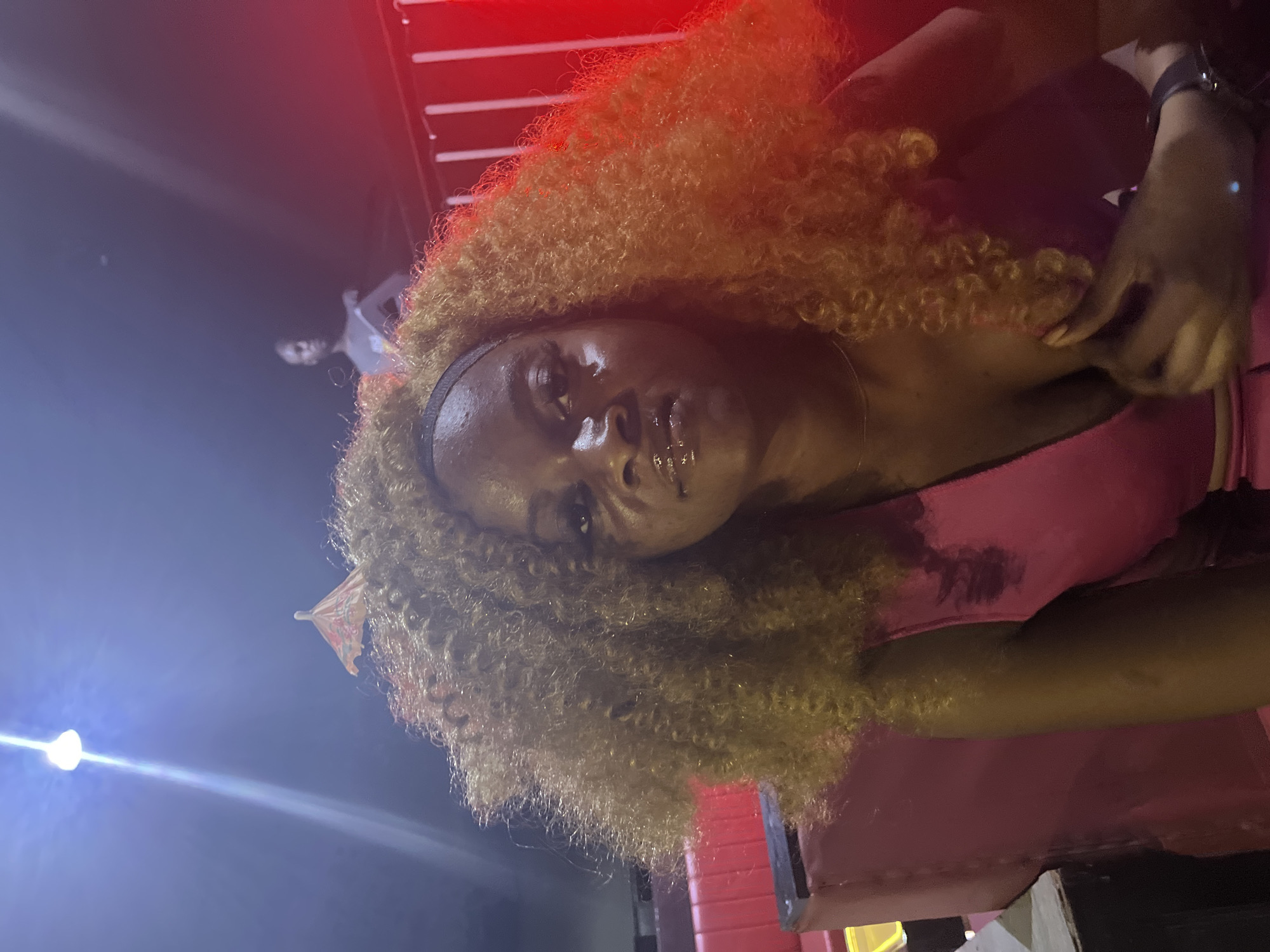 A woman with large, curly, ginger-colored hair is seated in a pink booth. She is wearing a shiny pink top and a thin necklace. A small umbrella is perched in her hair. In the background, a light fixture hangs from the ceiling, and another person is visible further back. The scene is illuminated by red lighting, suggesting a nighttime or dimly lit environment. The location is identified as Howayi, Nigeria.