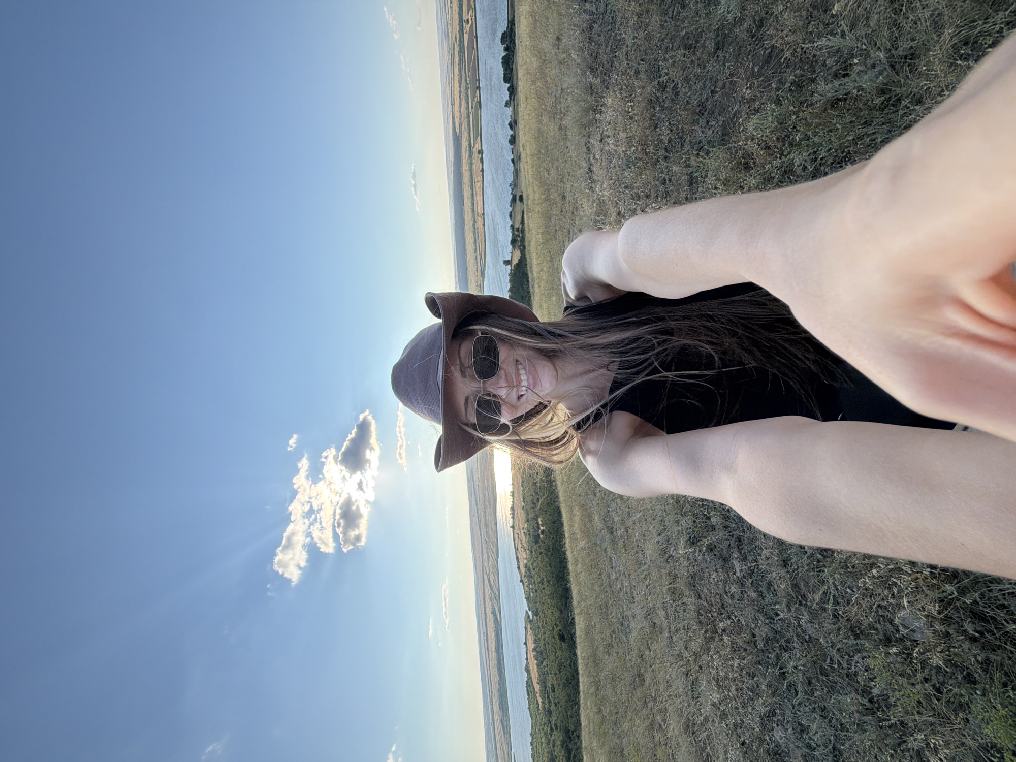 A selfie is taken by a woman wearing a brown cowboy hat and sunglasses, a black tank top, and a wide smile. She is standing on a grassy hill overlooking a wide river or lake with distant, sparsely vegetated shores. The sky is blue with scattered clouds and the sun is shining brightly. The woman's arms are extended forward, holding the camera. The environment appears to be rural, with dry grass covering the foreground and middle ground. The lighting suggests it is late afternoon or early evening. The location is the city of Gornyi Balyklei, Russia.