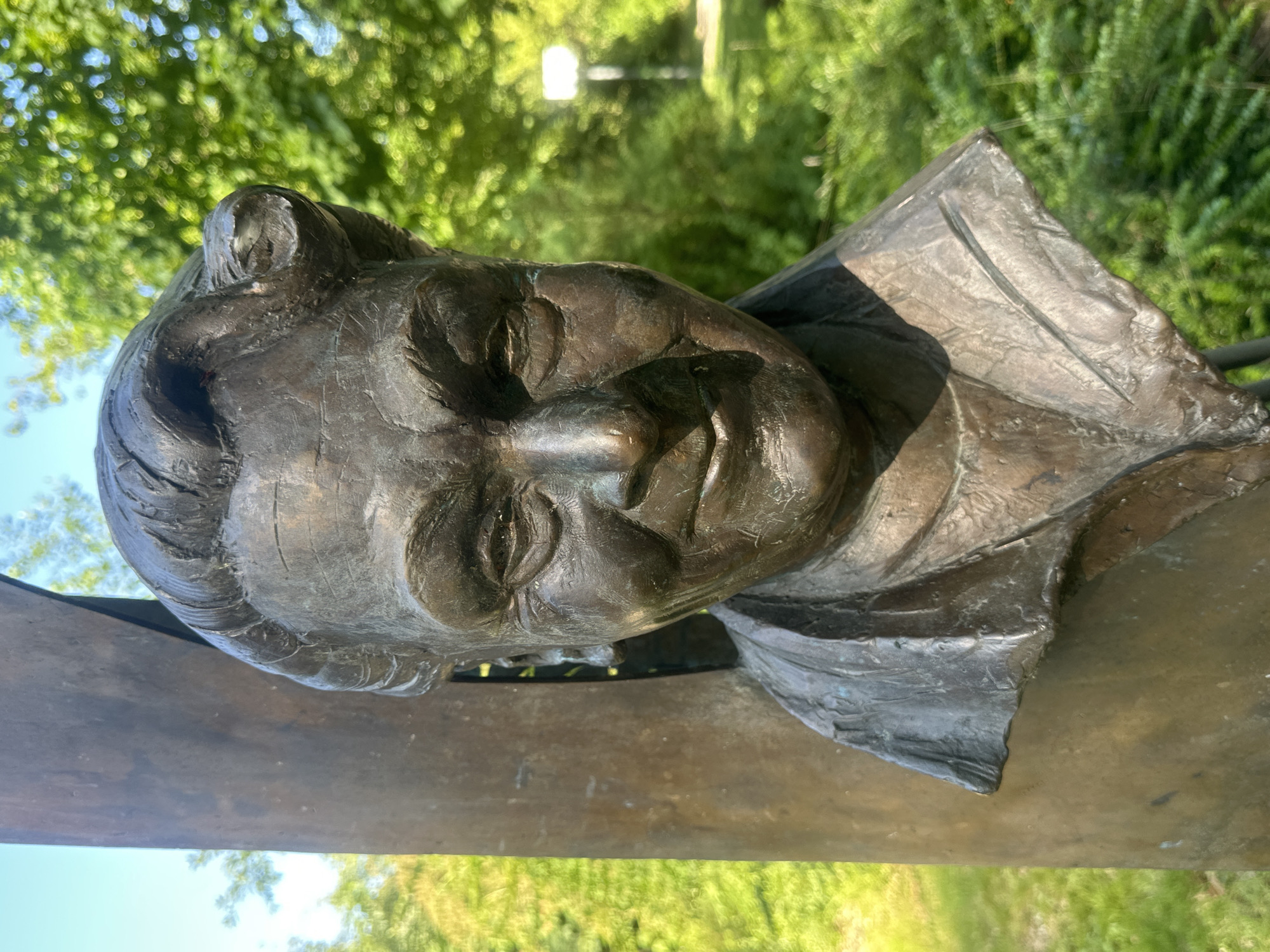 A close-up, eye-level, outdoor photograph captures a bronze bust of a woman positioned against a backdrop of lush green foliage and a clear blue sky. The bust is mounted on a vertical bronze column, which is partially visible on the left side of the frame. The woman's face is turned slightly to the left, and she has a warm, gentle smile. Her hair is styled in soft waves, pulled back from her face. Her clothing appears to be a collar and lapels, also rendered in bronze. The background is softly blurred, suggesting the bust is located in a park or garden setting. Sunlight illuminates the sculpture, creating highlights and shadows that accentuate its textured surface.