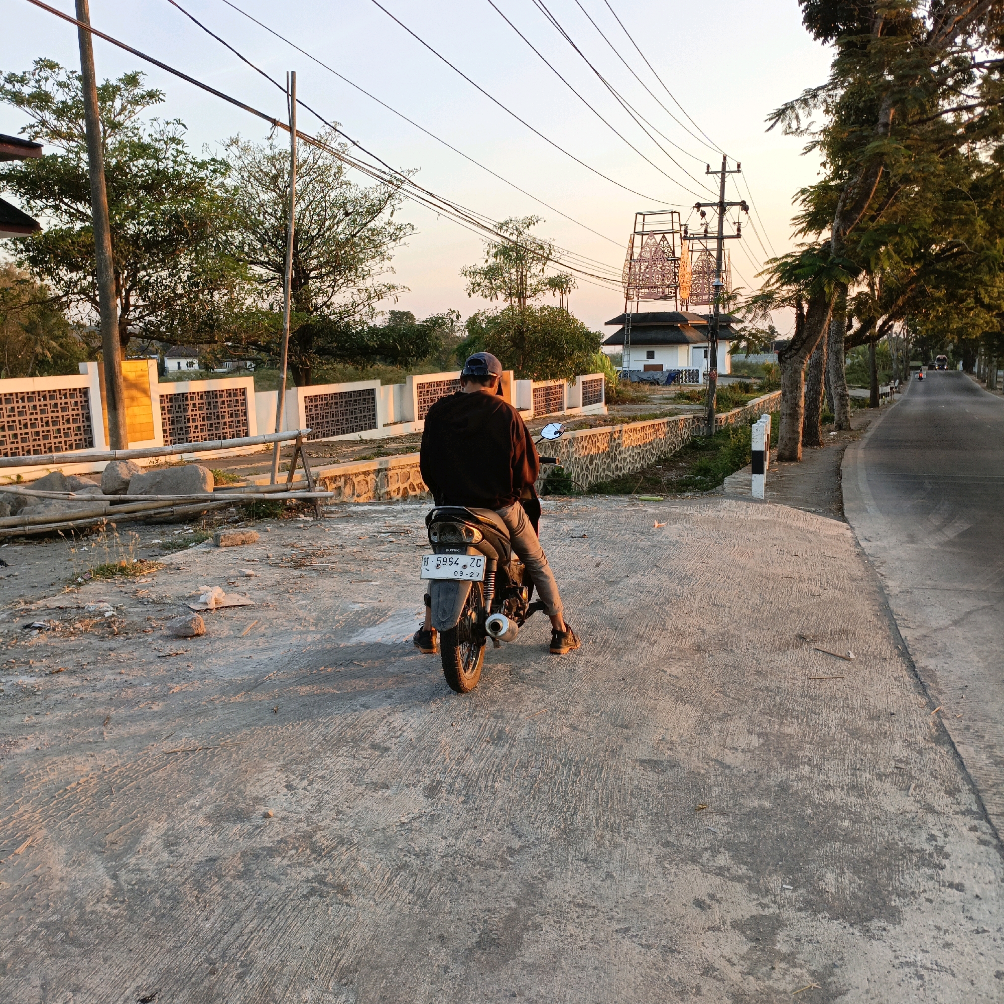 The image shows a person sitting on a motorcycle on a concrete road in Salatiga, Indonesia, at sunset. The person is wearing a black hooded sweatshirt, gray pants, a blue baseball cap, and brown shoes. The motorcycle is black with a white license plate that reads "B 2964 ZG 09-27". In the background, there is a white building with a decorative structure on top, a white fence with a patterned design, trees, and a utility pole with wires. The road is lined with trees on the right side. The sky is clear with a gradient of orange and yellow hues, indicating sunset. The concrete road is worn and shows tire marks. The environment appears to be rural or semi-urban.