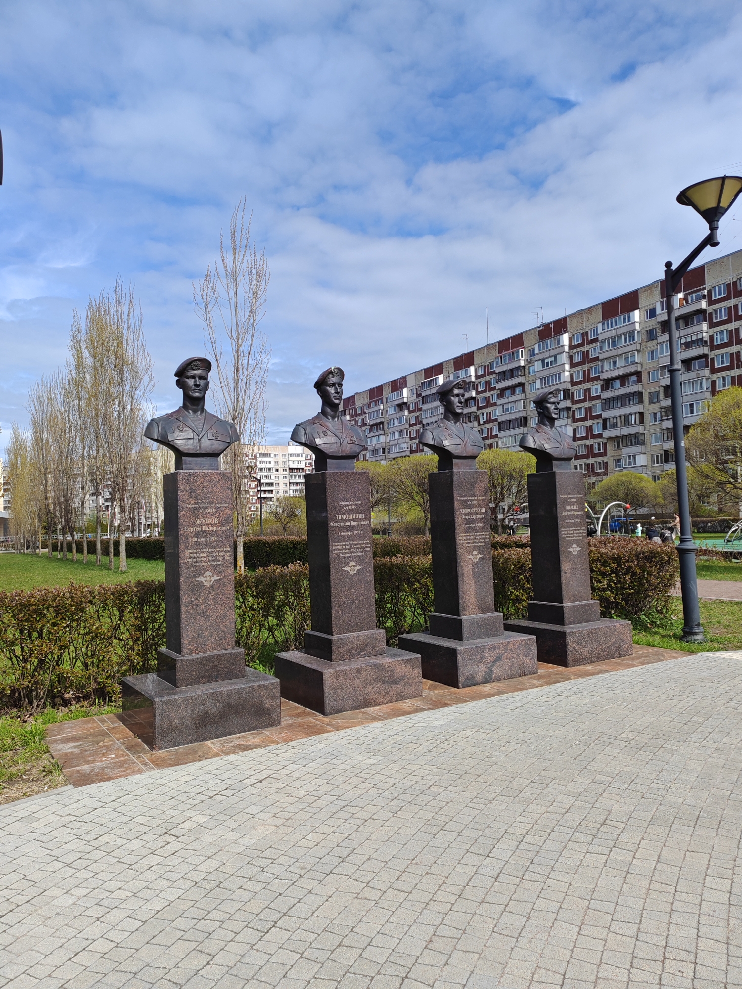 The image shows four bronze busts on dark-colored stone pedestals, arranged in a row along a paved walkway. These busts appear to be memorials to military figures, judging by their uniforms and the inscription on the pedestals.
Behind the monuments is a row of trimmed hedges and some bare trees. In the background, a large apartment building of light brick with brown accents rises several stories. A tall lamppost stands to the right of the last monument.
The scene appears to be a park or memorial garden. The location is identified as Saint Petersburg, Russia, based on the information provided.