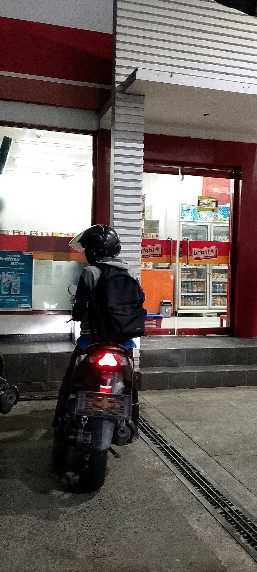 A person wearing a black helmet and a grey jacket is seated on a black scooter in front of a convenience store. The scooter's brake light is illuminated red. The store has glass doors with "bright*" logos and displays refrigerators stocked with beverages. The exterior of the store is painted red. A metal grate runs along the pavement in front of the store. The scene appears to be in the city of Bekasi, Indonesia.