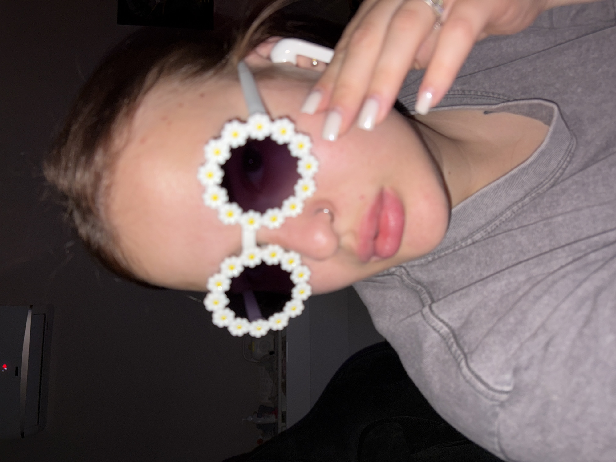 A close-up, slightly tilted image captures a person from the chest up, positioned against a dark background, likely indoors. The person's face is partially visible, featuring a nose piercing and slightly parted lips. They are wearing distinctive white daisy-shaped sunglasses with dark lenses, obscuring their eyes. A white Apple AirPods earbud is visible in their right ear.
The person's right hand is raised, with fingers partially covering their ear and temple area. Their fingernails are long and appear to have a light, neutral polish. A ring with a clear or white stone is visible on a finger of the raised hand. The person is wearing a gray long-sleeved garment, possibly a sweatshirt or hoodie, with visible stitching details. The overall lighting is dim, casting shadows and creating a soft, undefined background. To the left, a dark rectangular object with a small red light is faintly visible in the background, suggesting an electronic device. No discernible text is present in the image.