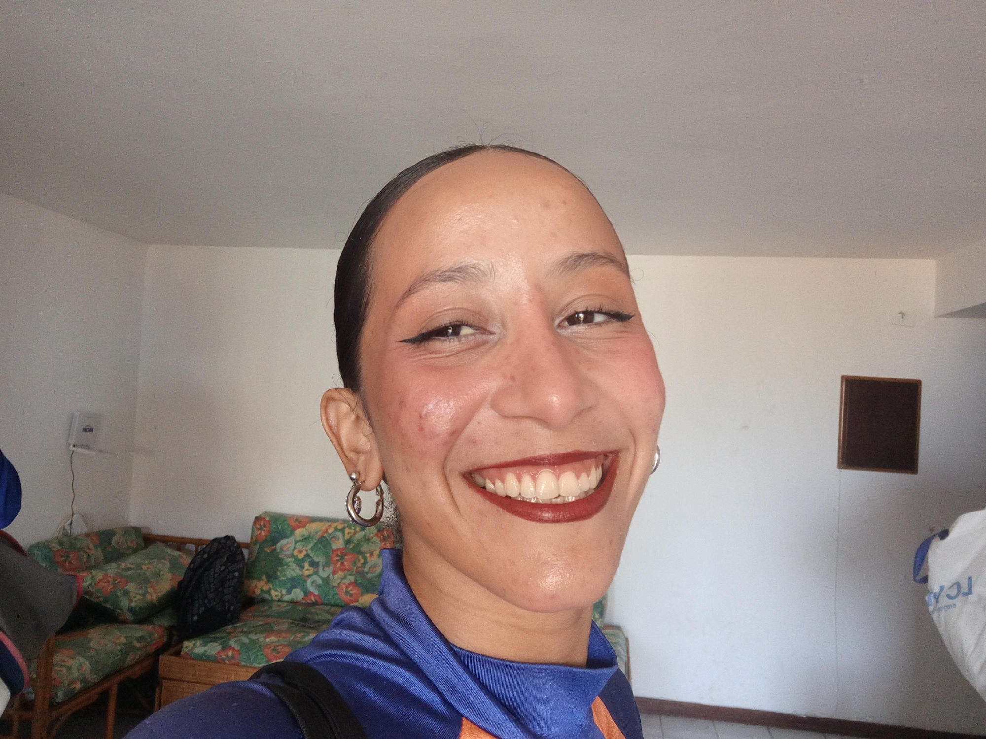 A woman is smiling into the camera. She is wearing a blue shirt with orange trim and hoop earrings. She has brown lipstick and eyeliner. A floral-patterned couch is visible in the background. A brown square object is attached to the wall. A white sack-like object stands in the corner. The location is likely indoors in Porlamar, Venezuela, based on other visual clues.