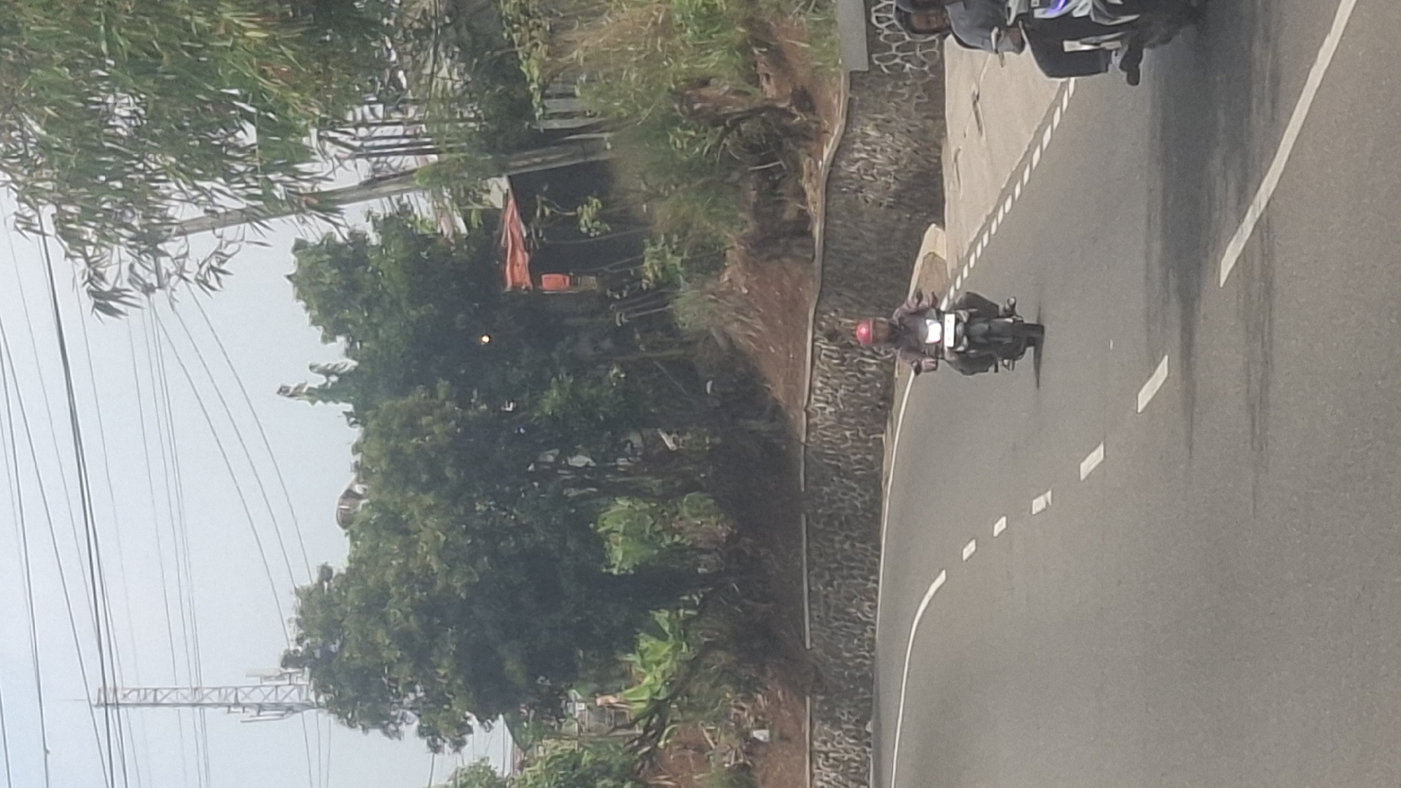 The image shows a view of a road with two motorcycles driving on it. The first motorcycle, closer to the foreground, has a single rider wearing a red helmet and a light-colored jacket. The motorcycle is dark-colored. The second motorcycle is further back and to the right, with two people on it, both wearing helmets. The road is paved with white lane markings. On the left side of the road, there is a retaining wall made of stone, and behind it, a grassy slope with trees and some structures. On the right side of the road, there is more vegetation and a fence. In the background, there is a telecommunications tower and a hazy sky. The scene appears to be in a rural or semi-rural area, possibly with some elevation given the slope on the side of the road.
