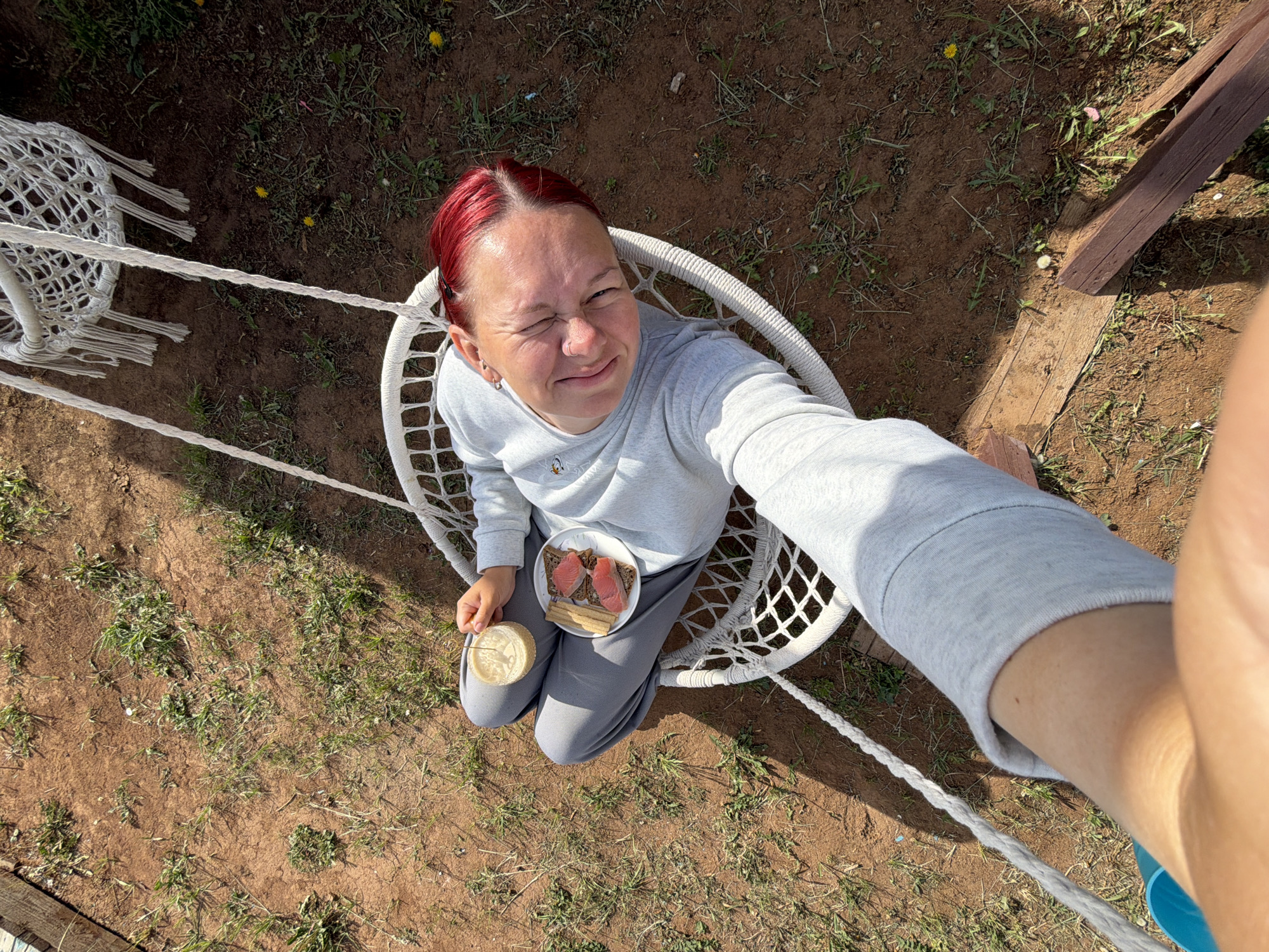 A high-angle selfie shows a woman with reddish-dyed hair, wearing a light gray sweatshirt and gray pants, sitting in a white macrame chair. She holds a small plate with bread and what appears to be salmon, and a jar, possibly containing a beverage or spread. She winks at the camera. The chair hangs from ropes. The background is a patch of earth with sparse grass and weeds. A section of a wooden structure is visible in the upper right corner. Another white macrame chair is partially visible in the upper left. The location is Chaikovsky, Russia, as evidenced by the photo's metadata.