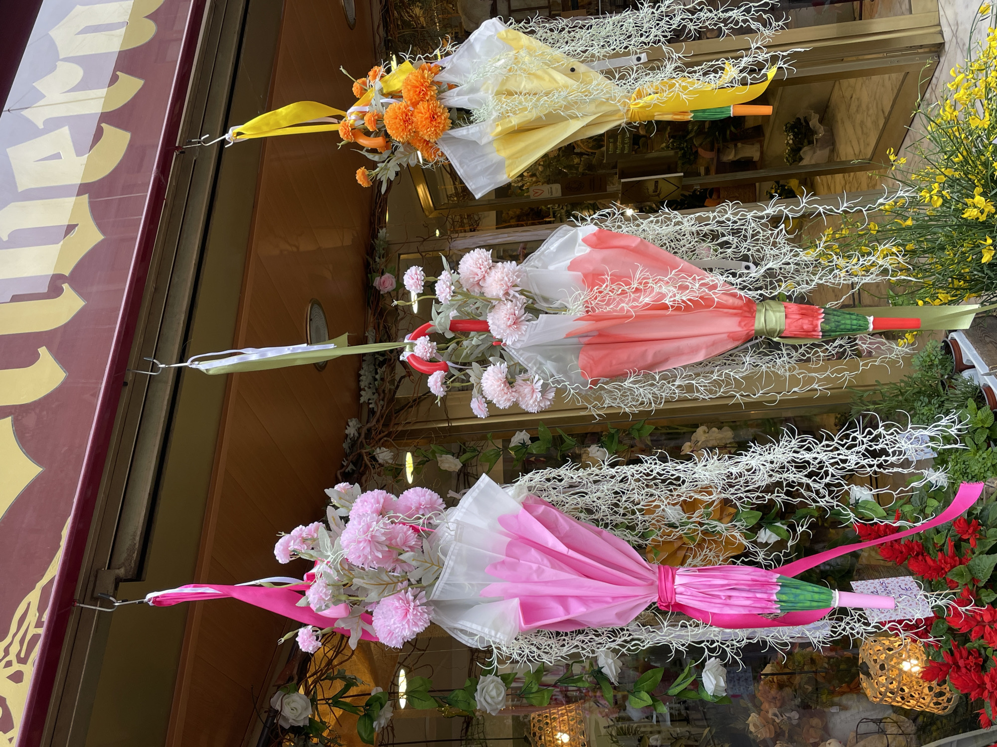 The image depicts a storefront in Terrassa, Spain, adorned with three decorated umbrellas. The umbrellas hang vertically, each a different color and embellished with flowers and white, twig-like decorations.
From top to bottom, the umbrellas are yellow with orange flowers, red with pink flowers, and pink with pink flowers. The umbrellas are all wrapped with ribbon, matching the color of the umbrellas, around the handles. The umbrellas are attached to the storefront awning with metal hooks.
The storefront behind the umbrellas is filled with various floral arrangements and decorative items, including yellow flowers, green plants, red flowers, and assorted decorations. The awning above the umbrellas is burgundy with gold lettering. The scene appears to be a floral shop or a shop selling decorated umbrellas.