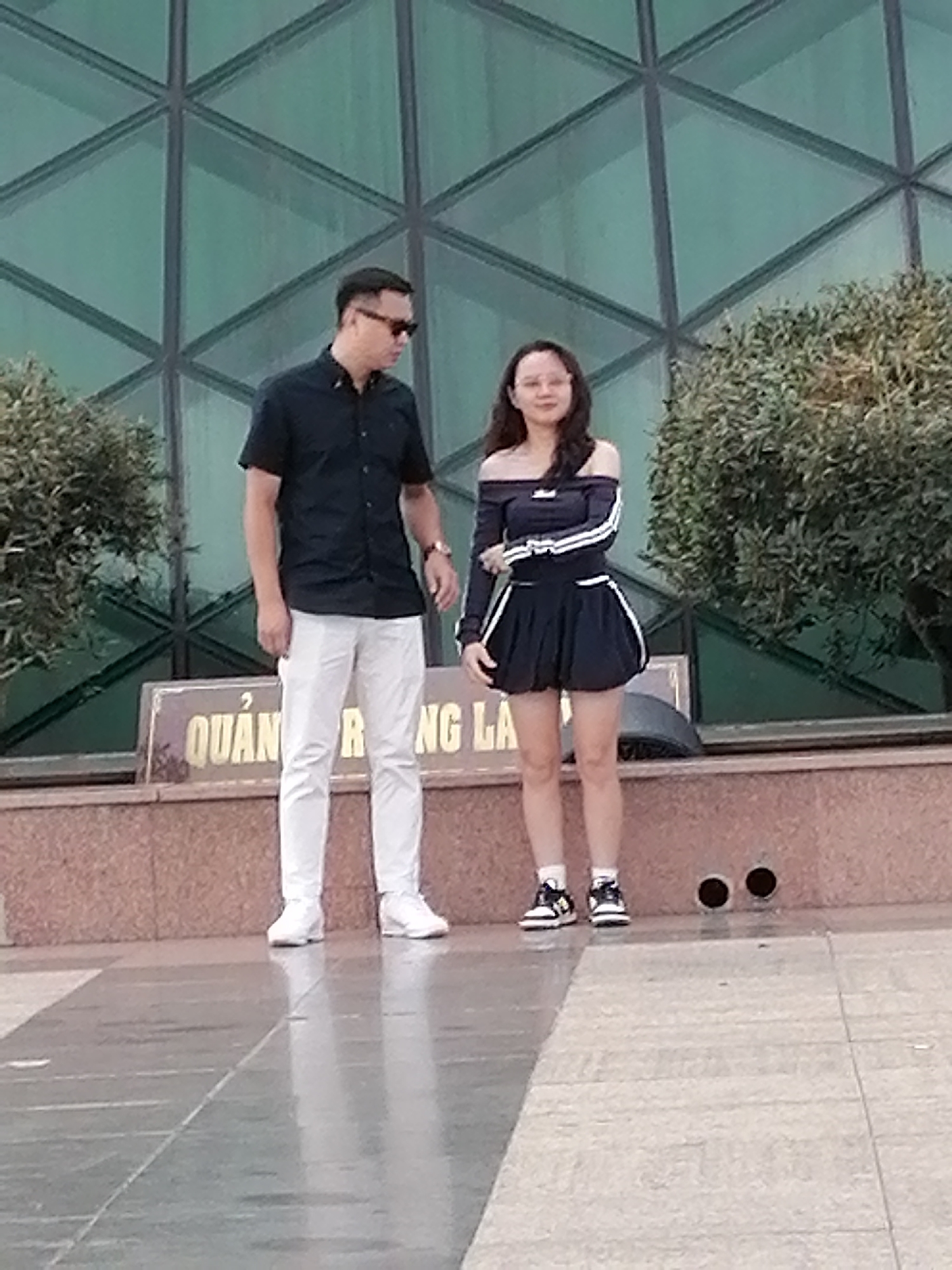 A man and a woman stand outdoors in front of a building with a green, geometric facade. The man, on the left, wears a black short-sleeved collared shirt, white pants, white sneakers, and sunglasses. The woman, on the right, wears an off-the-shoulder dark blue dress with white stripes on the sleeves and skirt, white socks, and black and white sneakers. A sign with Vietnamese text is visible behind them. The area appears to be a public space, possibly a plaza or walkway, with a paved surface and some greenery. The scene is likely in Da Lat, Vietnam, given the text on the sign.