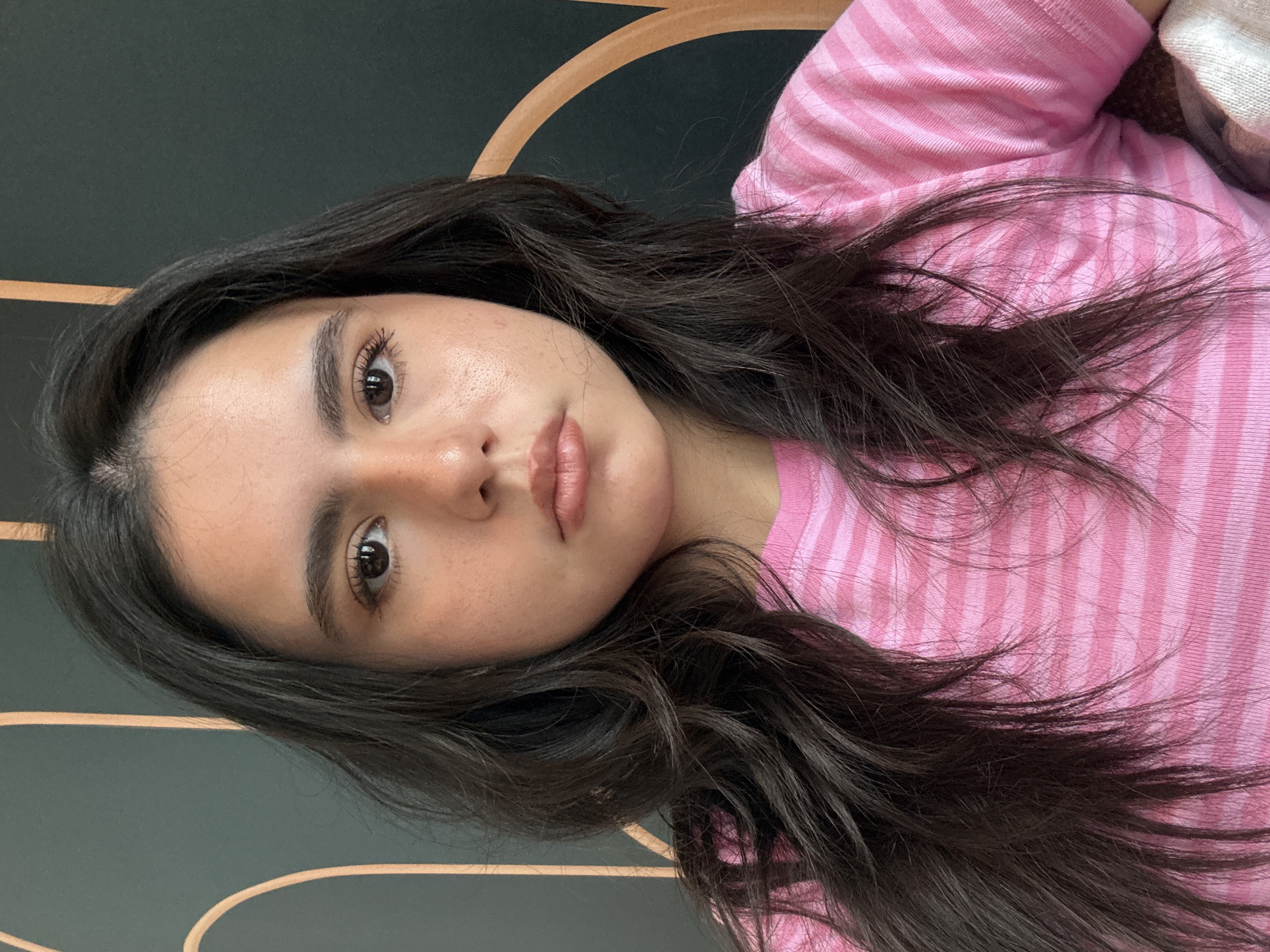 A young woman with dark hair and light brown eyes is lying down, wearing a pink and light pink striped shirt. The background appears to be a dark greenish-grey wall with tan or light brown decorative arches. The photograph seems to be taken indoors. There is insufficient data to determine if the photograph was taken in Austin, Texas.