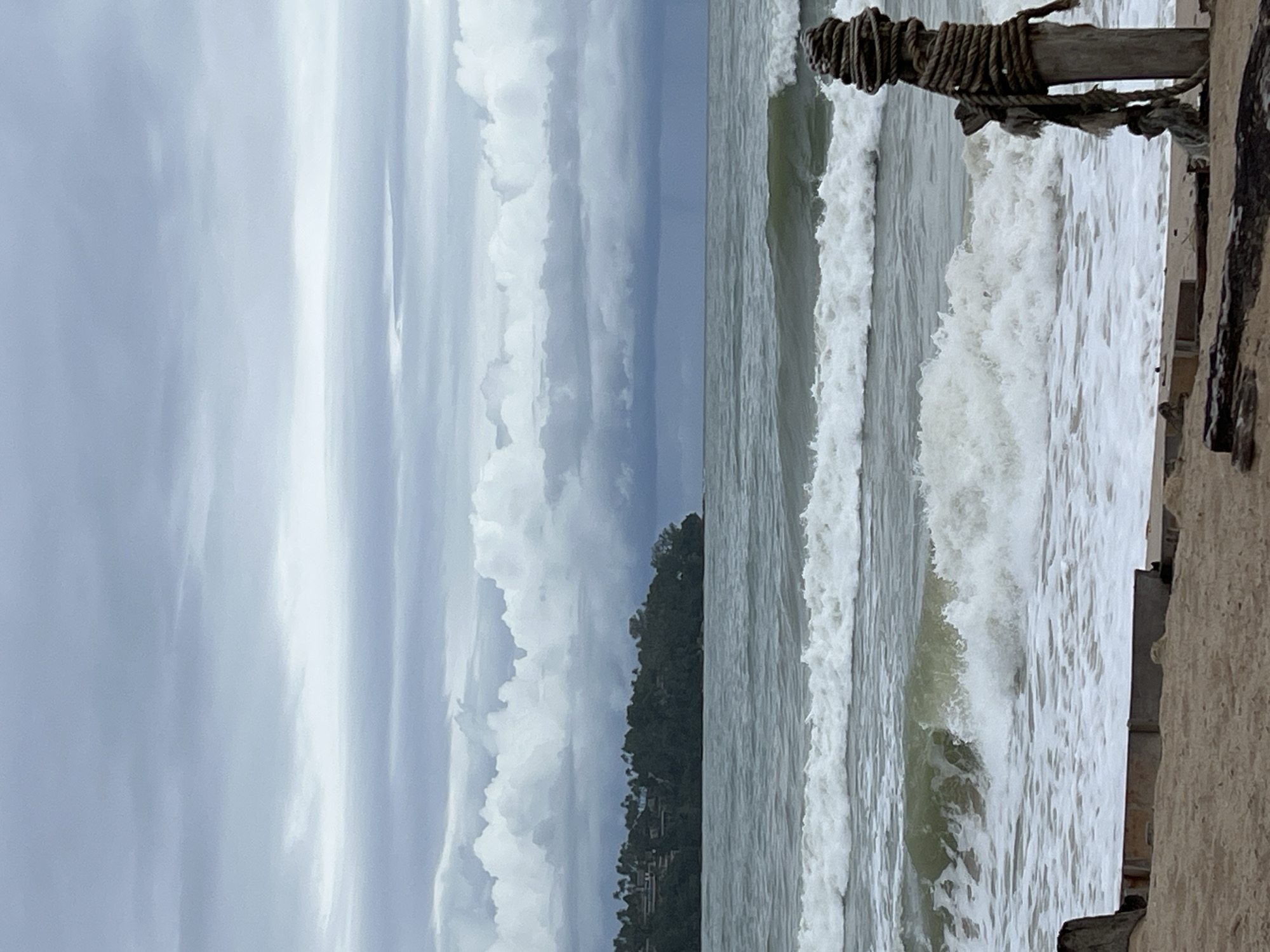 The image shows a choppy sea with several waves crashing on a sandy beach under a cloudy sky. In the distance, a lush green island with trees and buildings is visible. In the foreground, a weathered wooden post wrapped in thick rope stands on the beach, with some debris also present. The overall scene suggests a coastal environment, possibly during a period of overcast weather.