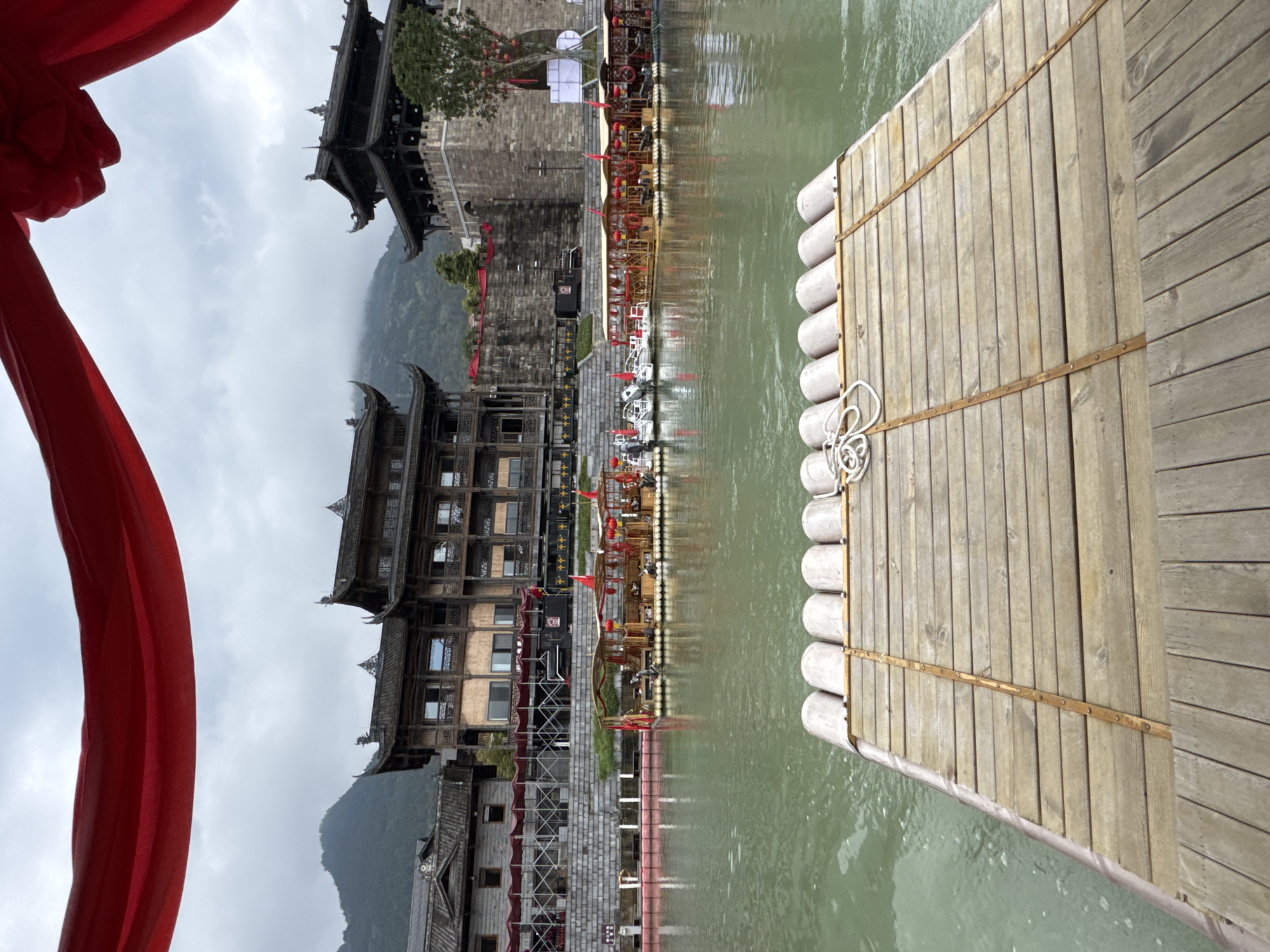 View from a wooden raft on the Zhuoshui River in Zhuoshui, China. The raft's wooden planks and rounded, flotation logs are visible in the foreground. A rope is tied in the middle of the raft. The murky river extends to the middle ground, reflecting the architecture on the shore. The architecture features traditional Chinese structures with ornate rooftops and wooden detailing. Red decorations adorn the buildings and bridges. A red cloth is visible on the left side of the image. Mountains are visible in the background under an overcast sky.