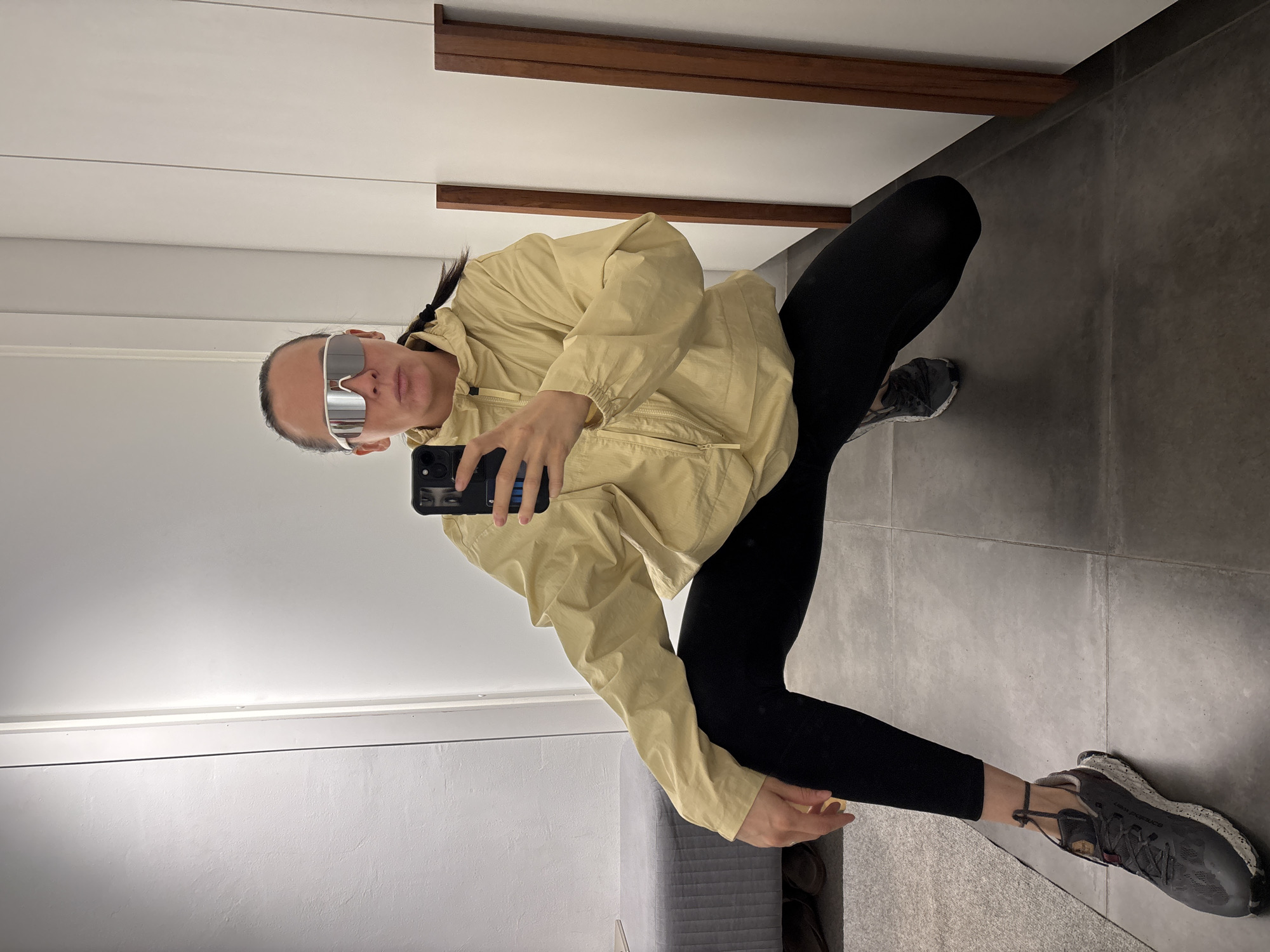 The image displays a full-body mirror selfie of a person crouching indoors, likely in Chaikovsky, Russia. The individual, who appears to be female, is captured in a deep squat or crouch, holding a black smartphone in her right hand as if taking the picture. She is wearing a light yellow or beige hooded windbreaker jacket, black leggings, and dark grey athletic shoes with speckled white soles. Prominent, large, reflective silver sunglasses cover her eyes, giving a sporty and somewhat obscured look. Her dark hair is styled in a braid, visible on the right side of her head.
The setting is a modern indoor space with clean lines. The floor is composed of large, dark grey rectangular tiles, with a section of lighter grey carpet visible on the left side. The background, as seen in the reflection, features a plain white wall or paneling, accented by two horizontal dark wooden strips or shelves in the upper half. A grey, possibly upholstered bench or ottoman is partially visible on the bottom left. The black smartphone has multiple camera lenses visible on its back. On the sole of the right shoe, the brand name "New Balance" is visible. The lighting suggests an indoor daytime scene, and the overall impression is one of casual, contemporary style.