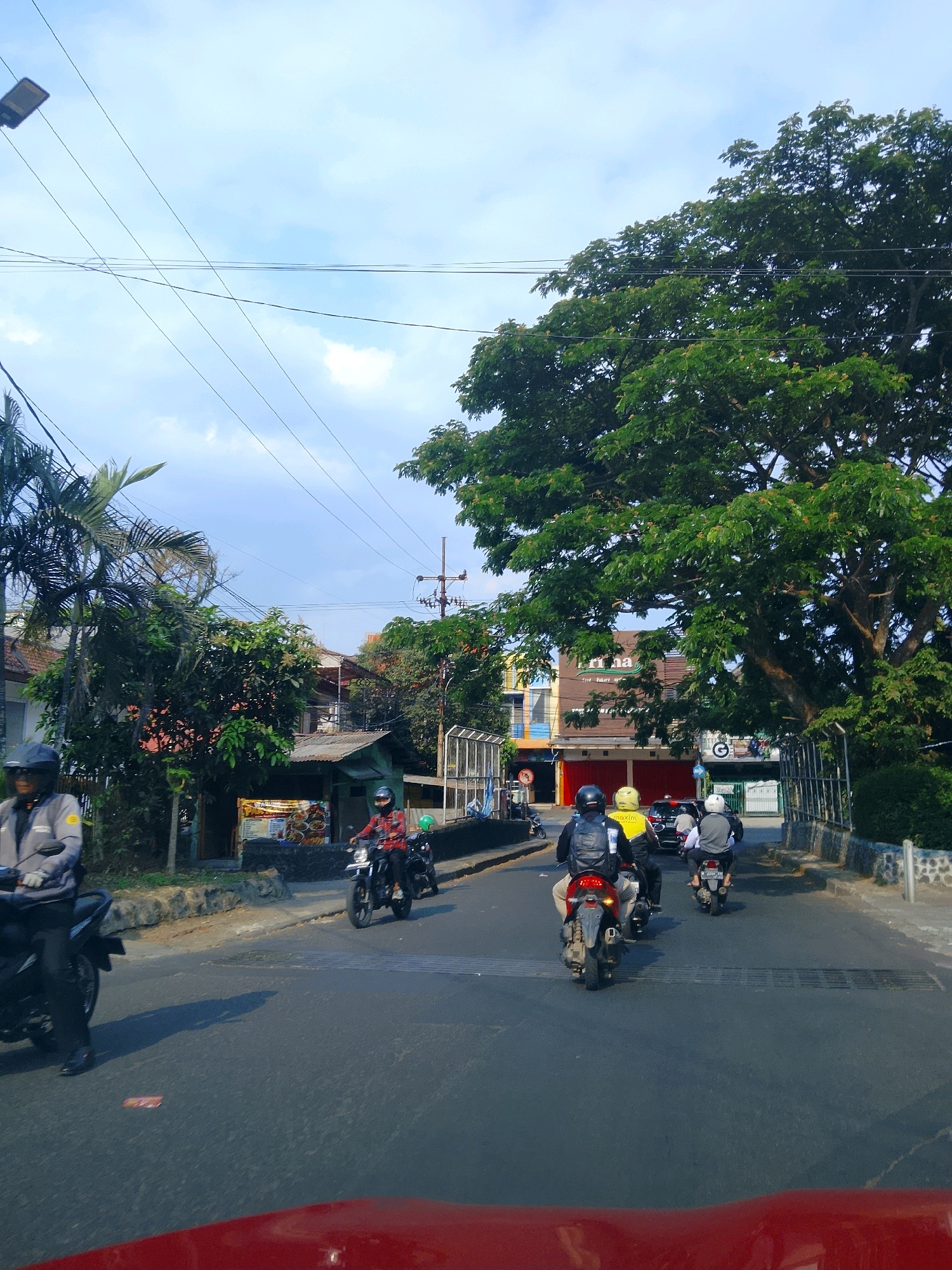 The image depicts a street scene in the city of Malang, Indonesia, under a clear sky with scattered clouds. Several motorcycles are traveling on the road. In the foreground, a red vehicle's hood is visible. On the left side of the road, a person wearing a gray jacket and a helmet rides a motorcycle. Further down the road, on the left, there are palm trees and buildings, including a small shop with food displayed. On the right side of the road, a large, leafy tree dominates the upper right portion of the frame. A group of motorcyclists are seen in the middle of the road, heading away from the viewer, with one rider wearing a bright yellow jacket. Buildings with signs are visible in the background, suggesting a commercial area. Power lines crisscross the sky, and a street lamp is visible on the left. The overall impression is that of a typical day on a street in Malang.