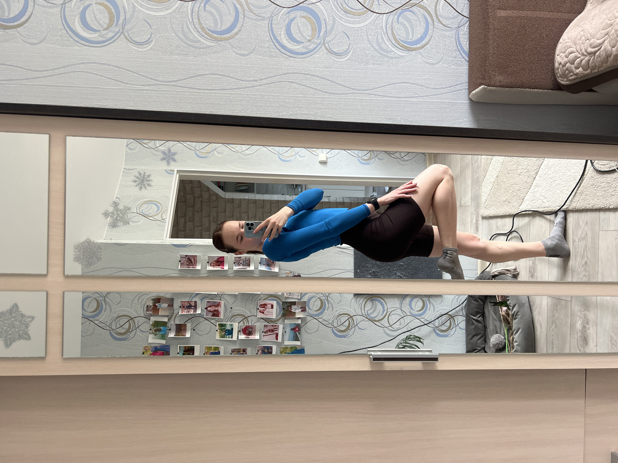 The image shows a woman in a blue long-sleeved shirt and dark shorts taking a selfie in front of a mirror. The mirror consists of multiple panels, with decorations such as snowflakes and photos attached to it. The surrounding environment is a room with patterned wallpaper, light-colored furniture, a brown rug, and a light-colored rug with a black cord running across it. The location is most likely an apartment or house in Chaikovsky, Russia.