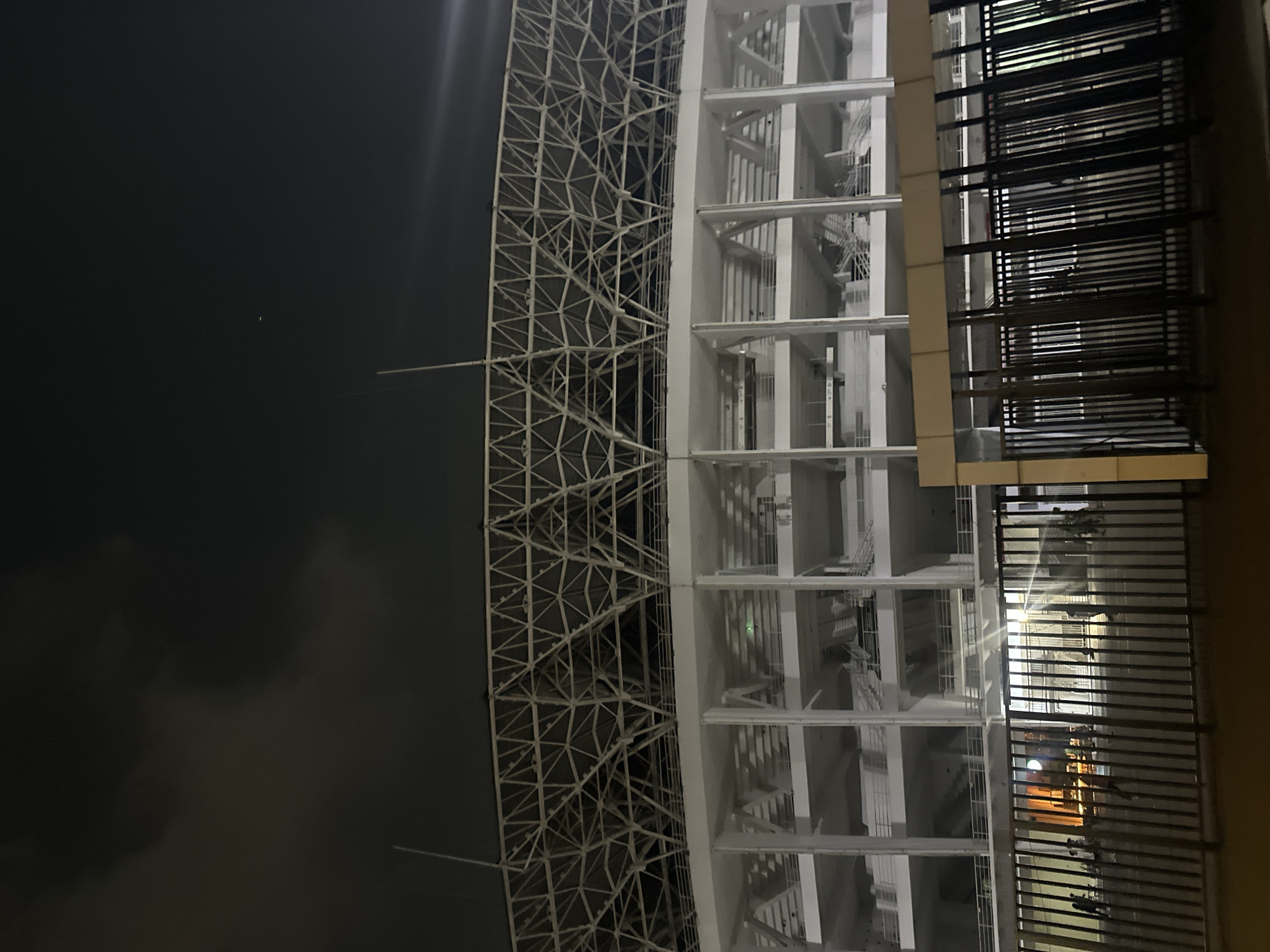 A nighttime view depicts the architecture of a building complex in Central Jakarta, Indonesia. The structure features a dense, web-like exterior design adjoining a multi-story, vertically-oriented building that includes balconies with dark-colored railings. The sky is dark and partially obscured by clouds. The image is rotated 90 degrees counter-clockwise.