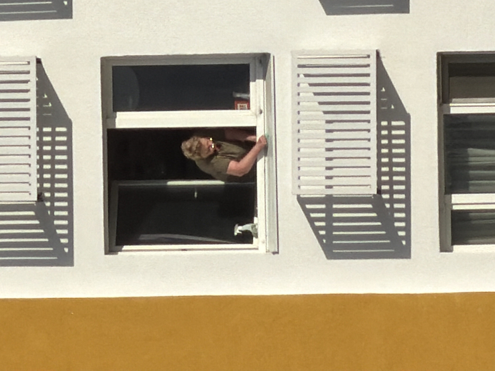 A person with short, blonde hair is leaning out of an open window. The person is wearing a dark green short-sleeved shirt. The window is surrounded by white walls. To the left and right of the window are white shutters. Below the white wall is a yellow section of the building. The scene appears to be outdoors in daylight. There is no indication of location being Khimki, Russia.