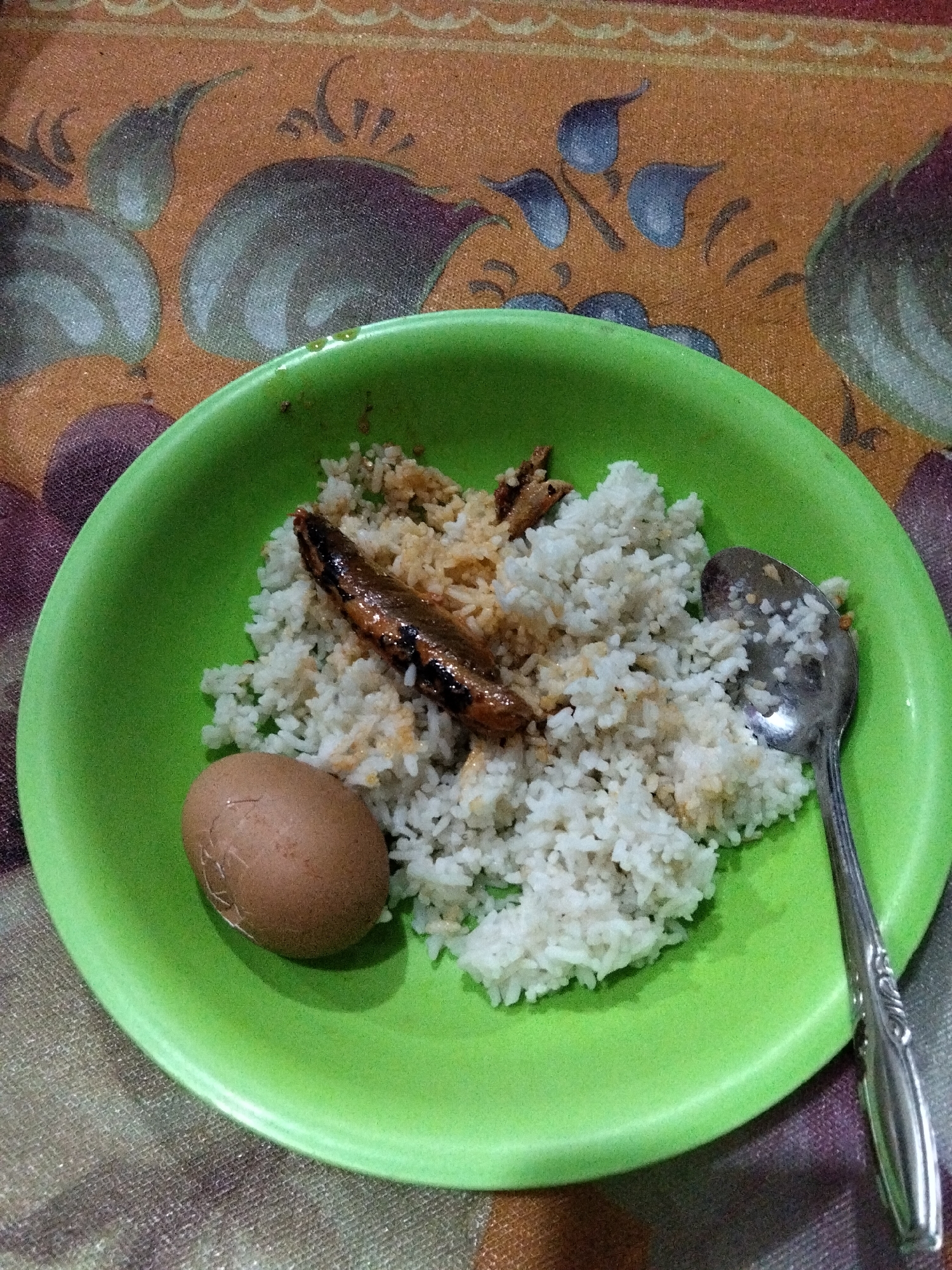 A plate of food sits on a patterned tablecloth. The plate is green and contains white rice, a hard-boiled egg, and what appears to be a fried fish. A silver spoon rests in the rice. The tablecloth has a pattern of stylized flowers and leaves in shades of brown, orange, and purple. The image appears to be taken in a domestic setting in the city of Benuaraja, Indonesia.