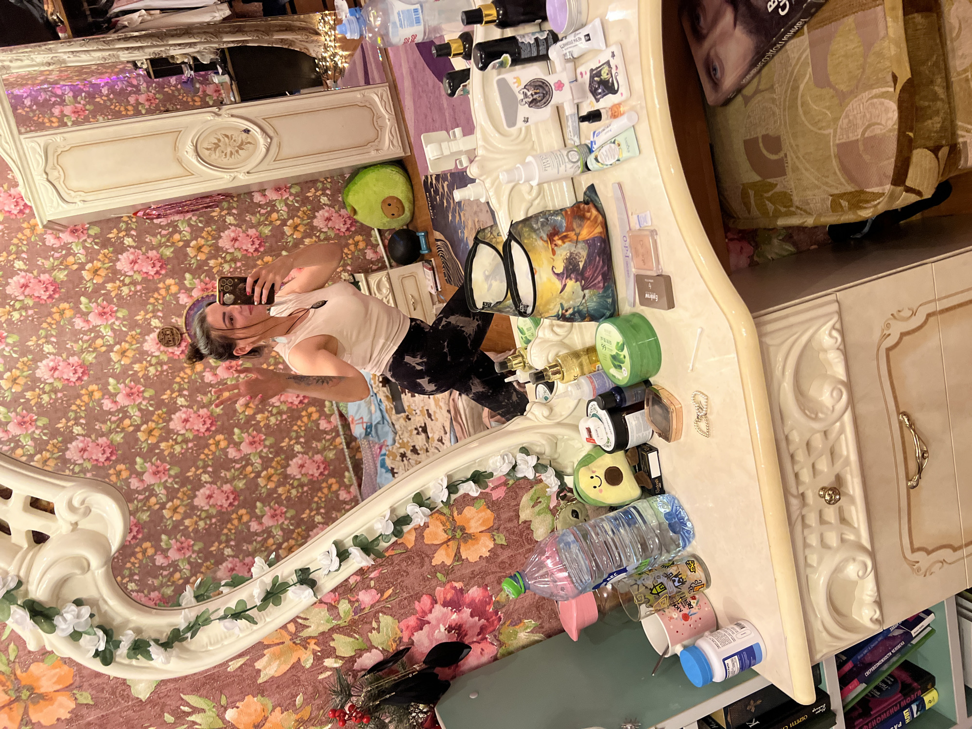 A young woman with dark hair tied in a bun, wearing a white tank top and black patterned leggings, takes a selfie in a mirror. She is standing in a room with floral wallpaper, in front of a vanity table cluttered with various cosmetic products, bottles, and bags. A large ornate mirror with white floral garlands is positioned on the vanity. To the right of the mirror, part of a white wardrobe is visible. The room appears to be a personal dressing area.