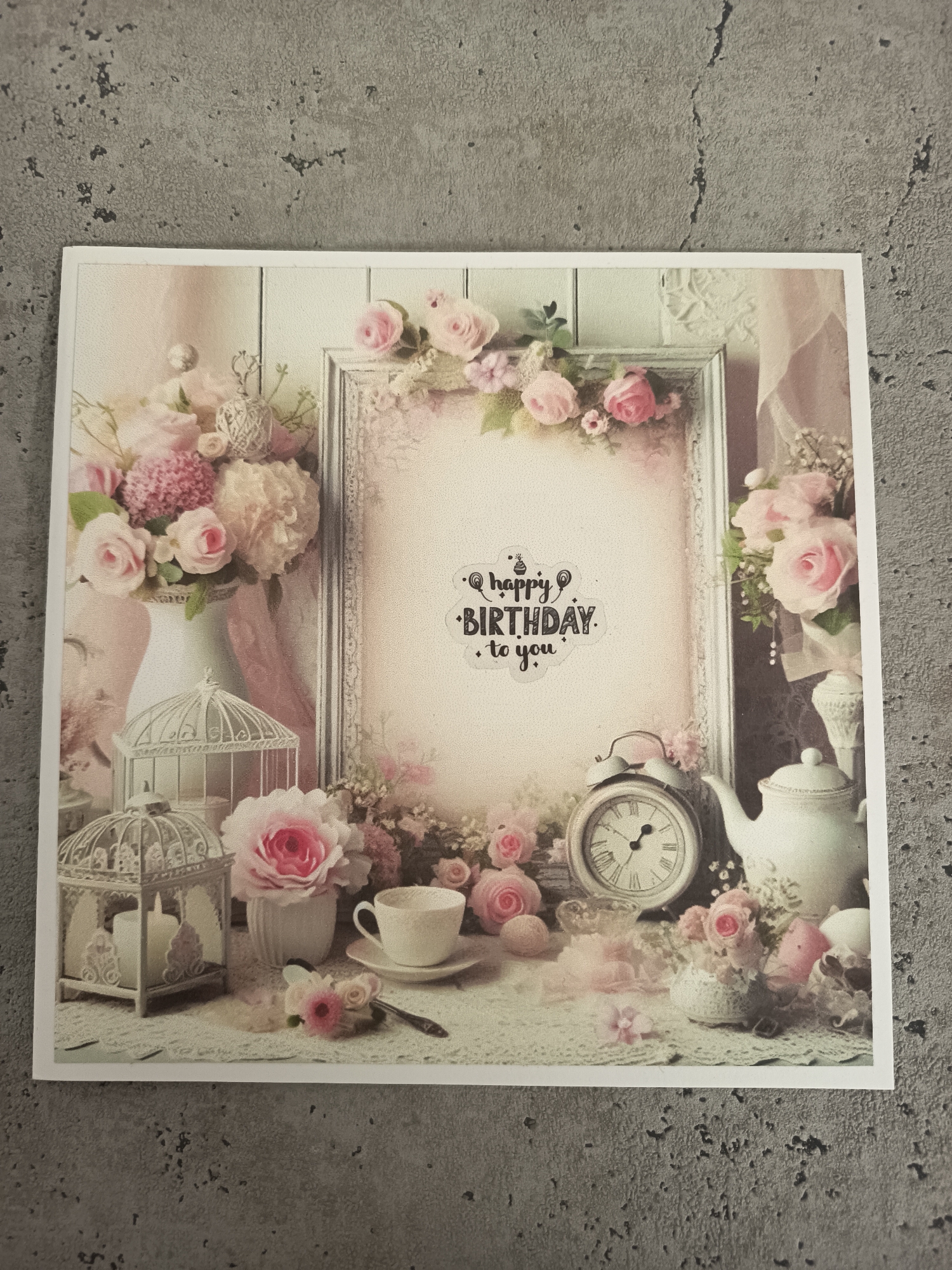 The image shows a birthday card with a romantic, vintage style. The card features a still life arrangement including pink roses, a birdcage, a teacup, a teapot, an alarm clock, and a picture frame with the words "Happy Birthday to you" written inside. The background of the card appears to be a wooden wall. The card is photographed on a gray, textured surface. There is no indication in the picture of Wangen bei Olten, Switzerland or any people.