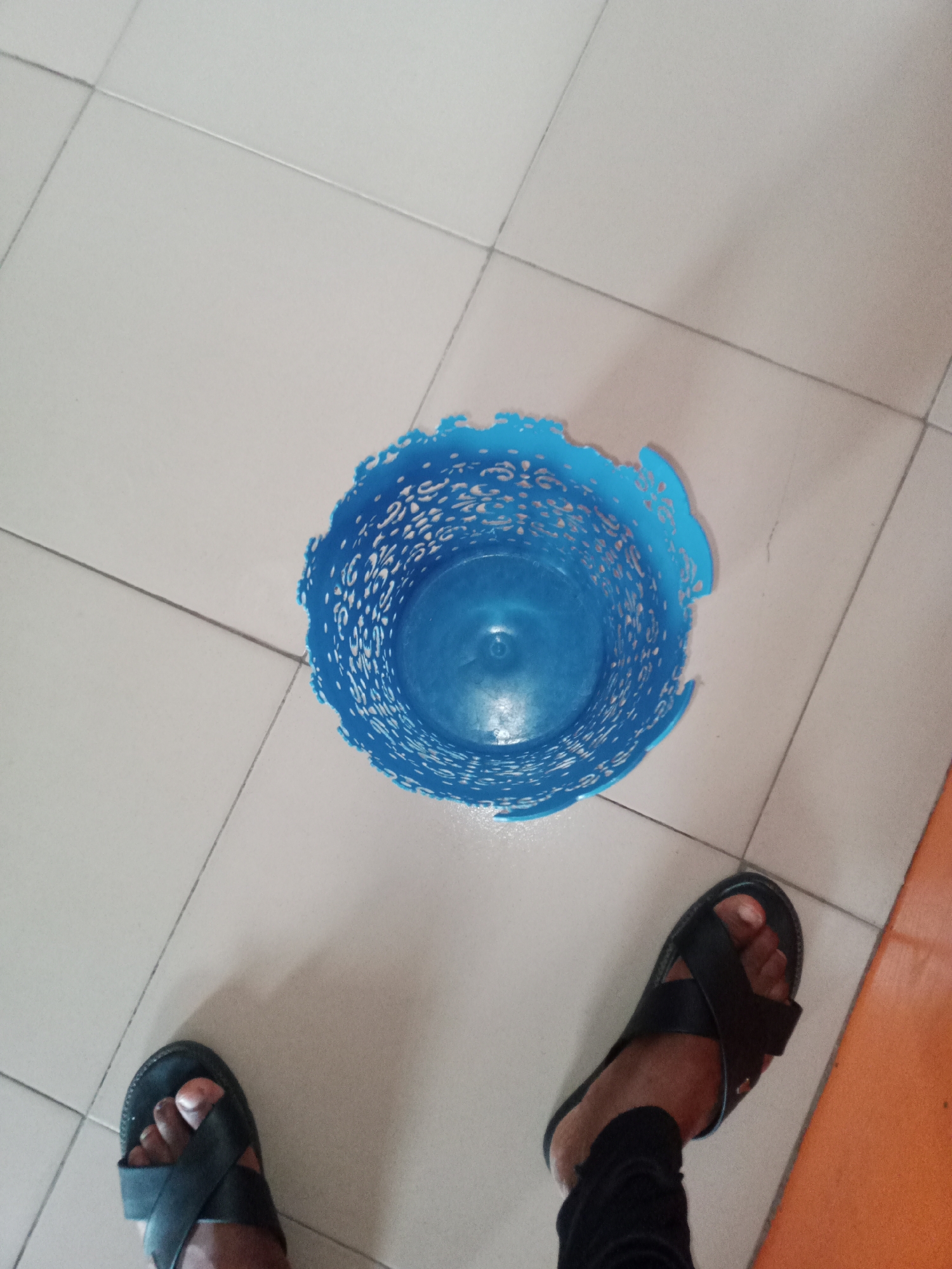 A pair of black cross-strap sandals are on a person's feet. The person is wearing black pants. The sandals and pants are on a light-colored tiled floor, with dark grout lines. In the center of the frame, a blue plastic waste bin with a decorative, perforated rim is on the floor. The waste bin is slightly damaged. The scene is indoors, likely in a room with a light-colored tiled floor and an orange wall visible on the right edge of the image. This could be a typical domestic or commercial setting in Jalingo, Nigeria.