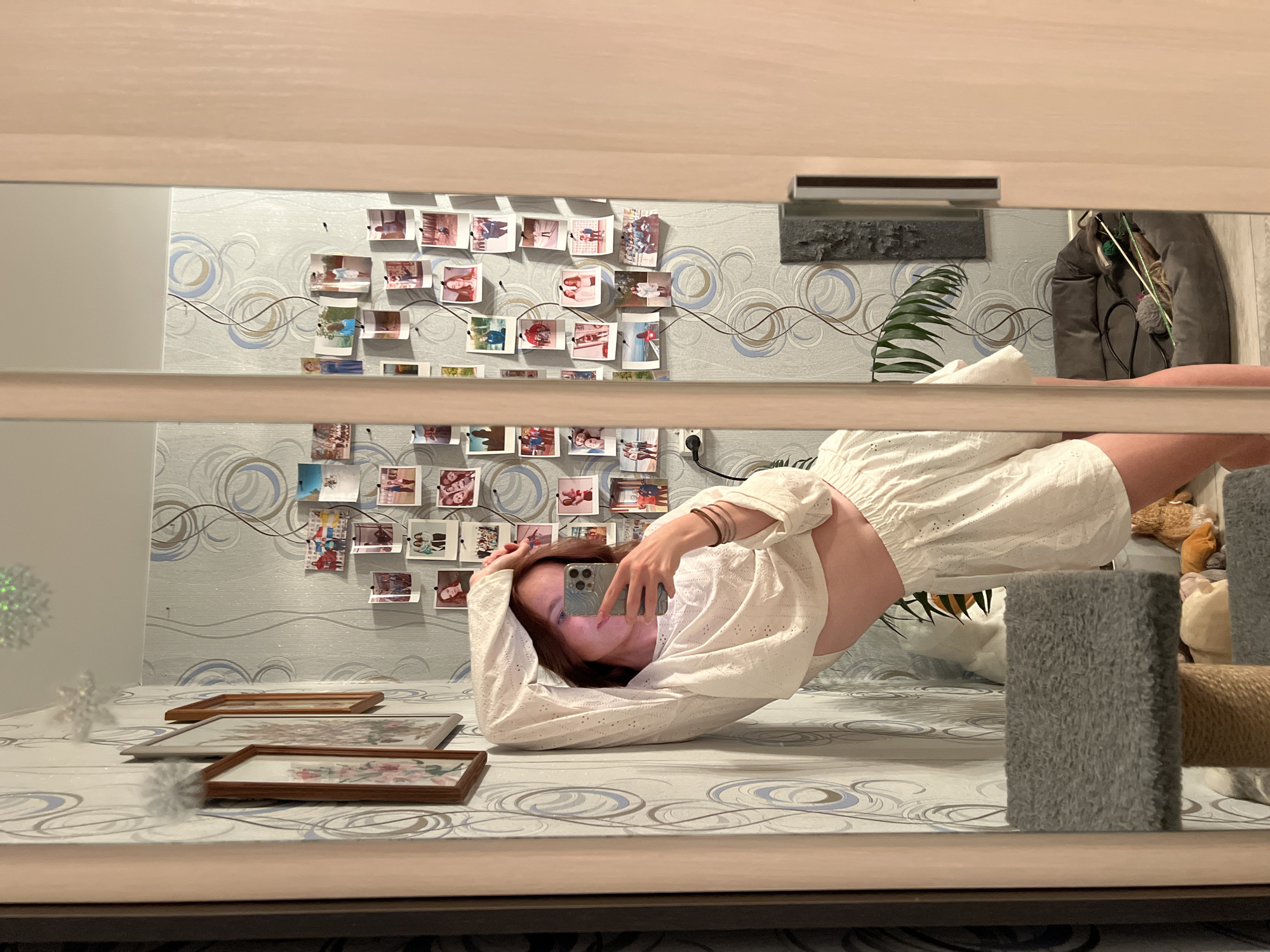 A reflection in a mirror shows a woman lying prone on a patterned surface, possibly a bed or mattress. She is wearing a white, eyelet-style shirt that exposes her midriff and a pair of matching white shorts. She holds a smartphone in her hand, presumably taking a selfie. In the background, the wall is decorated with a patterned wallpaper and a collection of photos pinned to it. Other objects in the reflection include framed pictures on the surface she is lying on, a plant, and what appears to be a cat tree or post. The location is Chaikovsky, Russia.