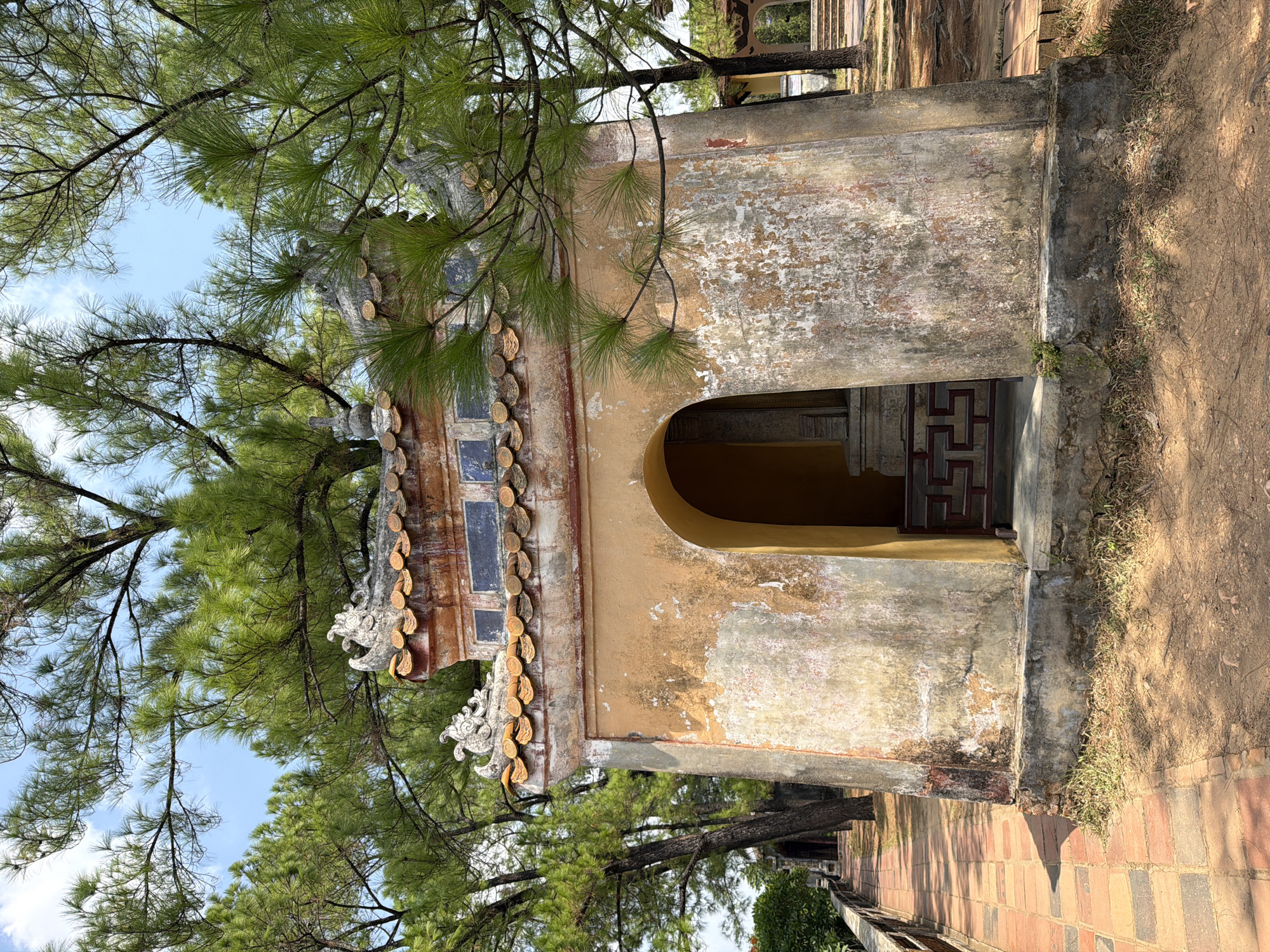 The image shows a weathered, yellow-ochre colored building with an arched entrance, set amidst trees. The building has a tiled roof with decorative sculptures. A brick pathway lined with a metal railing leads to the left of the frame, where other similar structures can be seen in the distance. The ground is covered in sand and dirt with scattered pine needles, and sparse grass grows at the base of the building. Sunlight filters through the canopy of the trees, casting shadows on the ground. The building appears to be a historical or religious structure, likely part of a larger complex. The location is identified as Kim Long Ward, Vietnam. There are no people visible in the image.