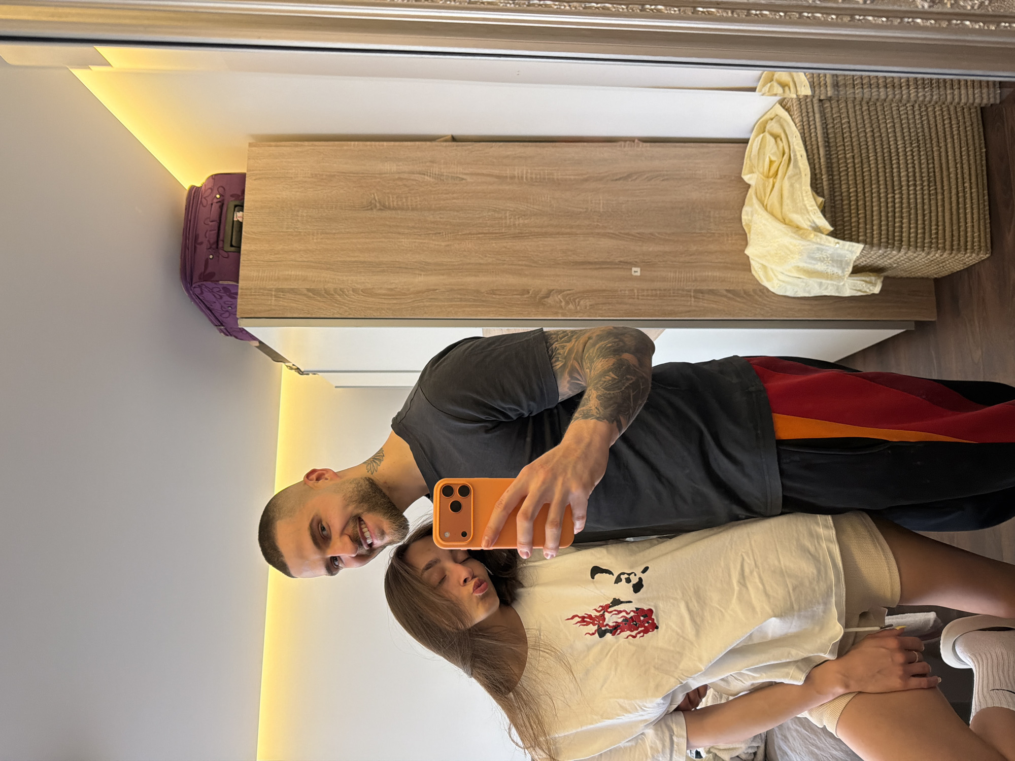 A man and a woman are posing for a selfie in what appears to be a bedroom. The man, with a shaved head and tattoos on his arm, is holding an orange phone, presumably taking the picture. He is wearing a dark gray t-shirt and black pants with red and orange accents. The woman, with long brown hair, is wearing a white t-shirt with a cartoon graphic and light-colored shorts. She is making a kiss face toward the camera. The background includes a light gray wall, a wooden cabinet, a wicker basket, and a purple suitcase. The reflection in the mirror suggests the location is likely in a modern apartment. While not definitively confirmed, given the language spoken and architectural style, this could potentially be in Sofia, Bulgaria.