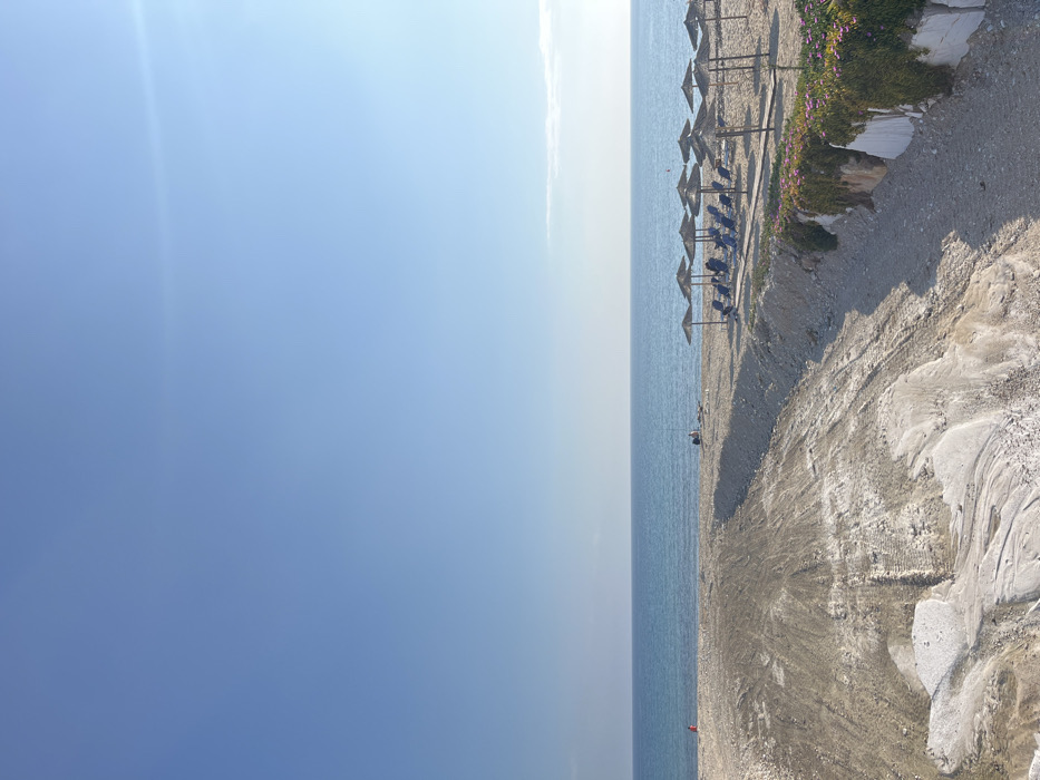 The image shows a view of a beach in Limenaria, Greece (latitude: 40.626, longitude: 24.573). The scene is captured from a slightly elevated vantage point, looking down towards the shoreline. The beach is mostly empty, with only a few people visible in the distance, sitting on lounge chairs beneath beach umbrellas. The sand is light-colored, interspersed with patches of gravel. There are several rows of lounge chairs and umbrellas lined up along the beach. In the foreground, a path leads down to the beach, with a small patch of greenery on the right side. The sky is clear and blue.