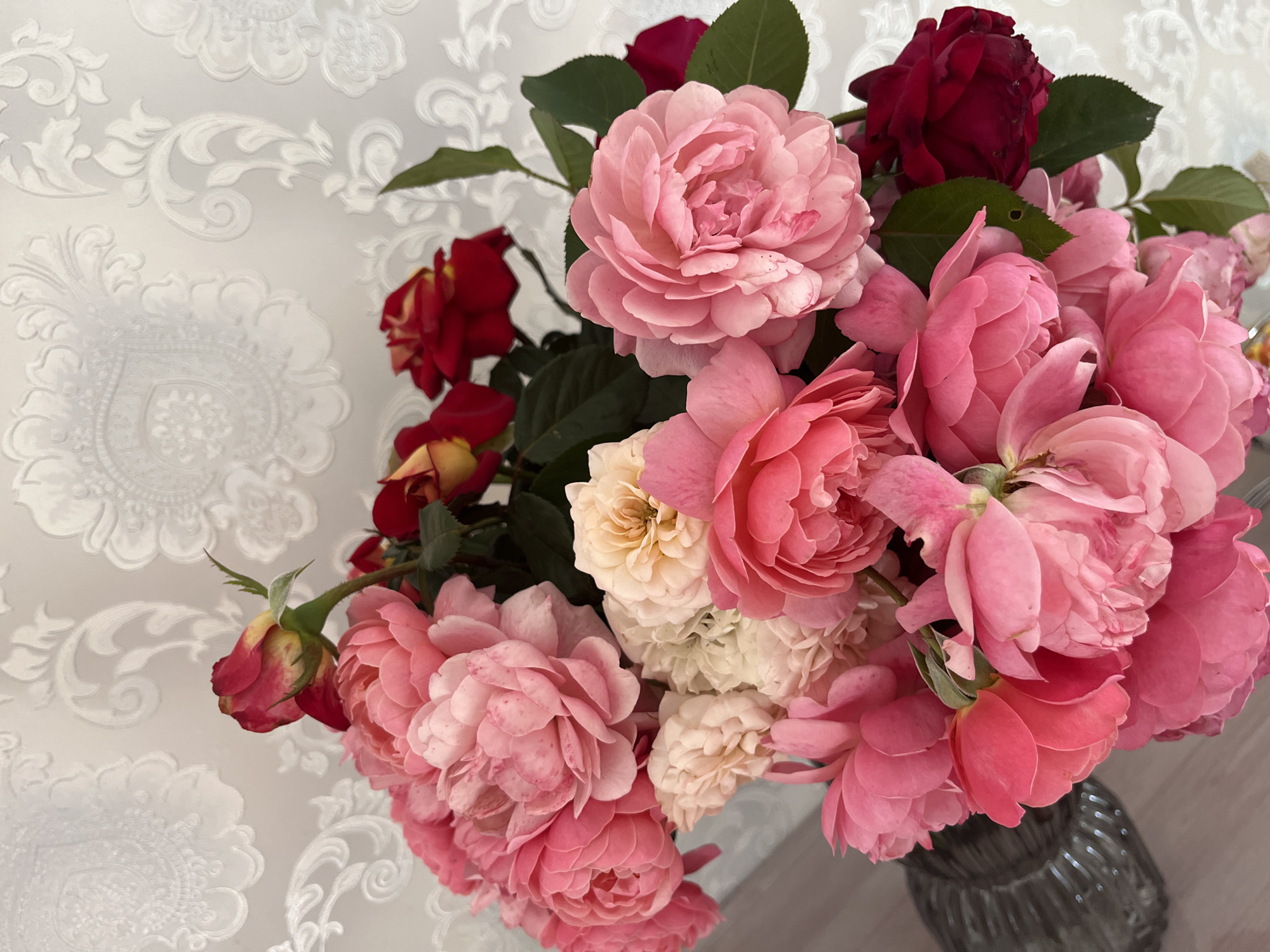 A bouquet of roses in shades of pink, cream, and dark red is displayed against a textured white wallpaper with an embossed floral pattern. The roses vary in bloom stage, with some fully open and others in bud. Green leaves are visible throughout the bouquet. The bottom left of the image shows a portion of a dark, textured vase. The scene appears to be indoors, likely in a domestic setting, given the wallpaper. The lighting is soft, suggesting natural or diffused artificial light. There are no people or animals visible in the image.