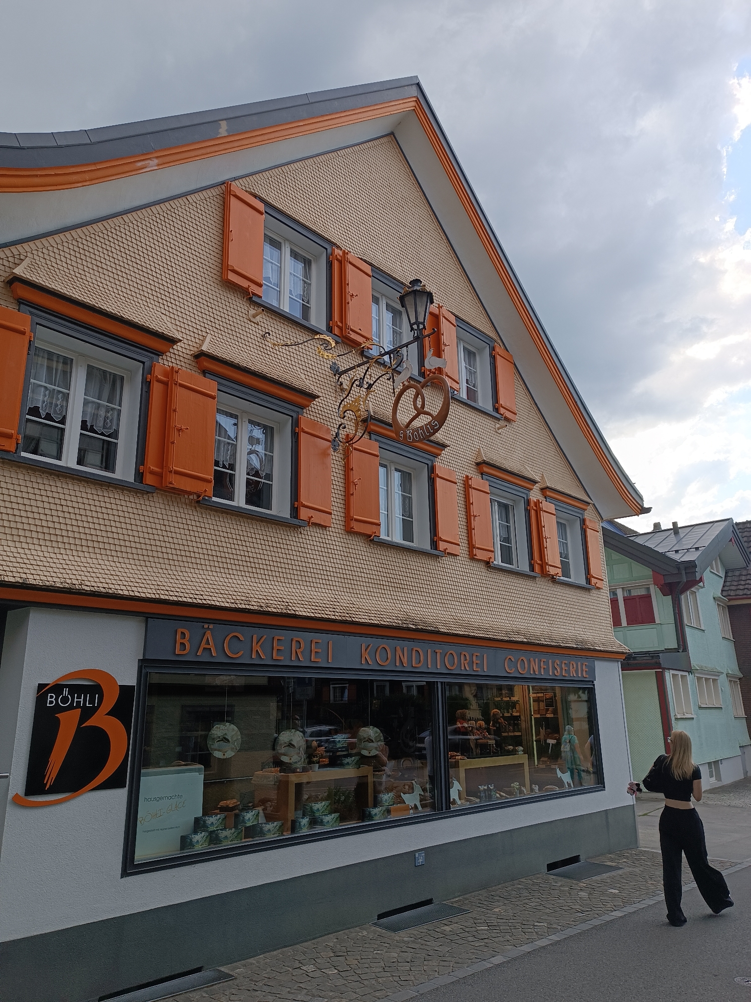 The image captures a street view of the exterior of a bakery and confectionary shop named "BÖHLI Bäckerei Konditorei Confiserie" located in the city of Appenzell, Switzerland. The building features traditional Swiss architecture with a tiled facade and bright orange shutters on its windows. A decorative sign with a pretzel motif hangs above the shop entrance. A young woman dressed in black, with blonde hair, is walking away from the camera on the paved sidewalk in front of the shop. The sky is overcast with visible clouds. The street is lined with cobblestones, and another building with a light green facade is visible to the right of the bakery.