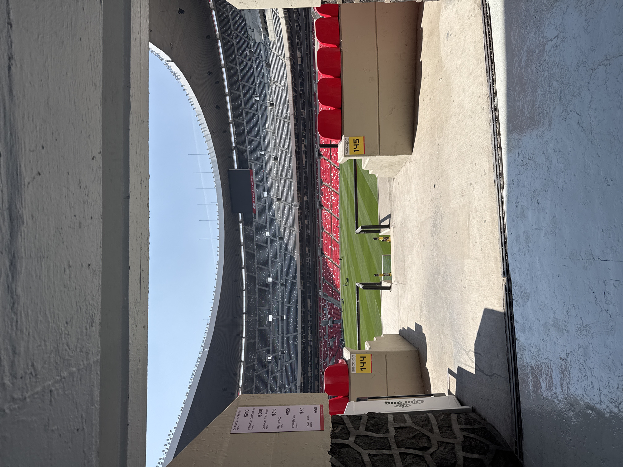 The image captures a view of Estadio Azteca in Mexico City, Mexico, a large stadium known for hosting soccer matches and other events. The perspective is from an elevated, enclosed passageway overlooking the field and seating areas. The stadium itself is massive, with tiers of seats in shades of gray and red that stretch upward. The green playing field is visible, with some maintenance equipment on it.
On the right side of the frame, there is a concrete structure that separates the passageway from the seating area. The numbers "145" are marked on it. Similarly, on the left side, there is another concrete structure with the number "144" and a sign showing prices for various drinks, including beer, soda, and water. The sign lists brands like Corona and Michelob.