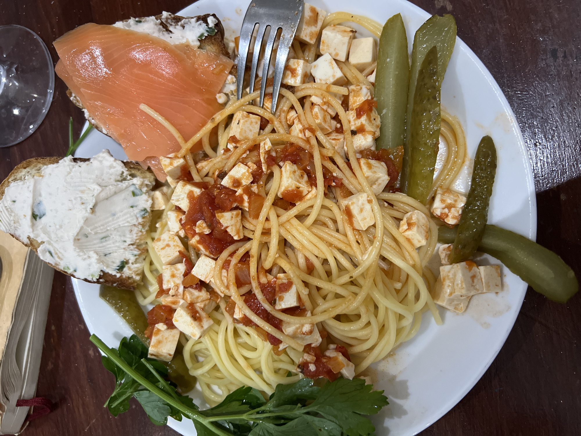 A plate of food is centered in the frame. The plate contains spaghetti with cubes of a white food item topped with red sauce, several pickles, and two slices of bread, each topped with different spreads: one is white, and the other has a salmon-colored topping. A fork is inserted into the spaghetti. Green leafy herbs are also on the plate. The plate rests on a dark surface. A wine glass is partially visible on the left, along with a book. The location is not specified.